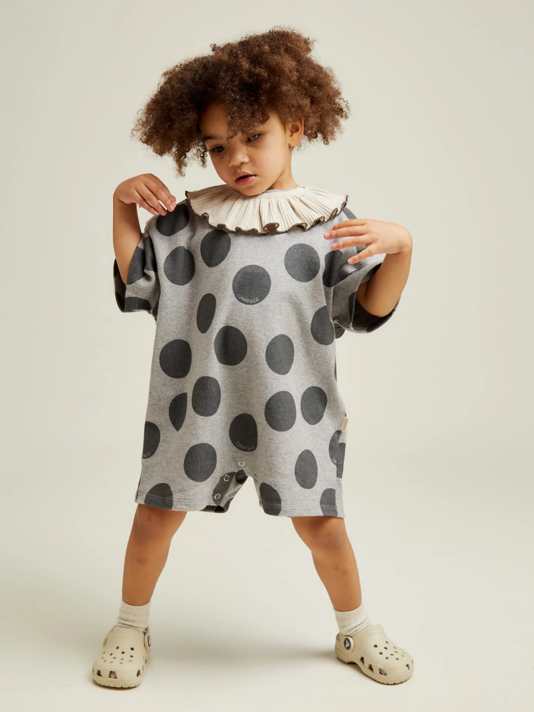 A young child with curly hair stands against a plain background, wearing the OPAL MARL SPOT ROMPER—gray with large black polka dots and a ruffled collar—paired with light-colored clogs.