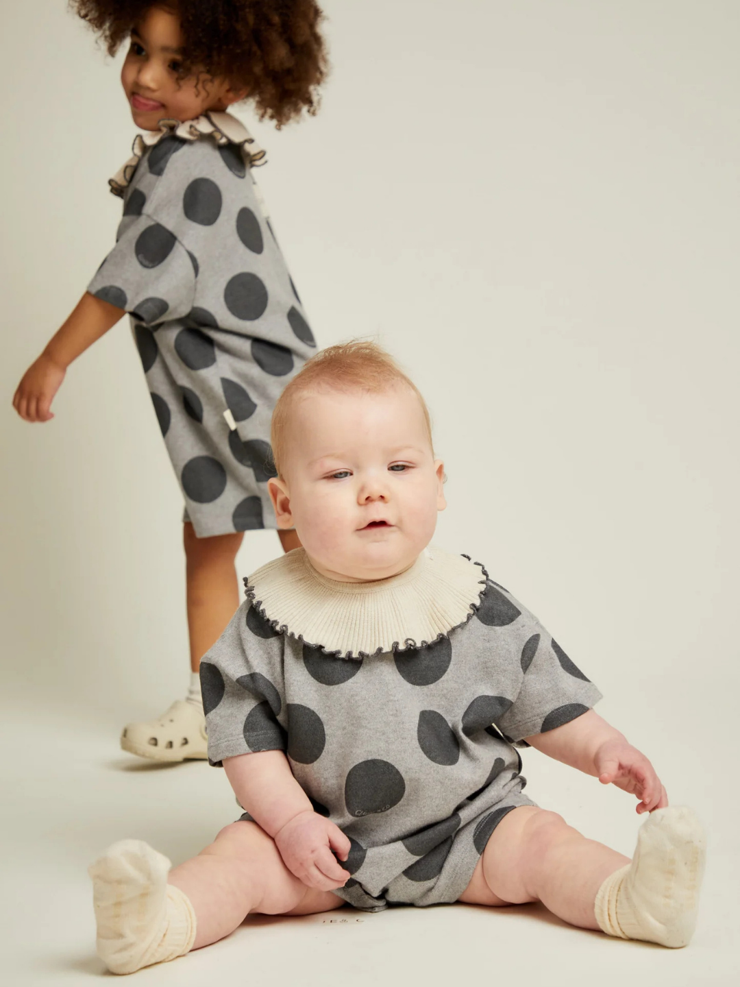 A baby wears the OPAL MARL SPOT ROMPER with large black polka dots and a ruffled collar, while a toddler in a matching outfit stands in the background.