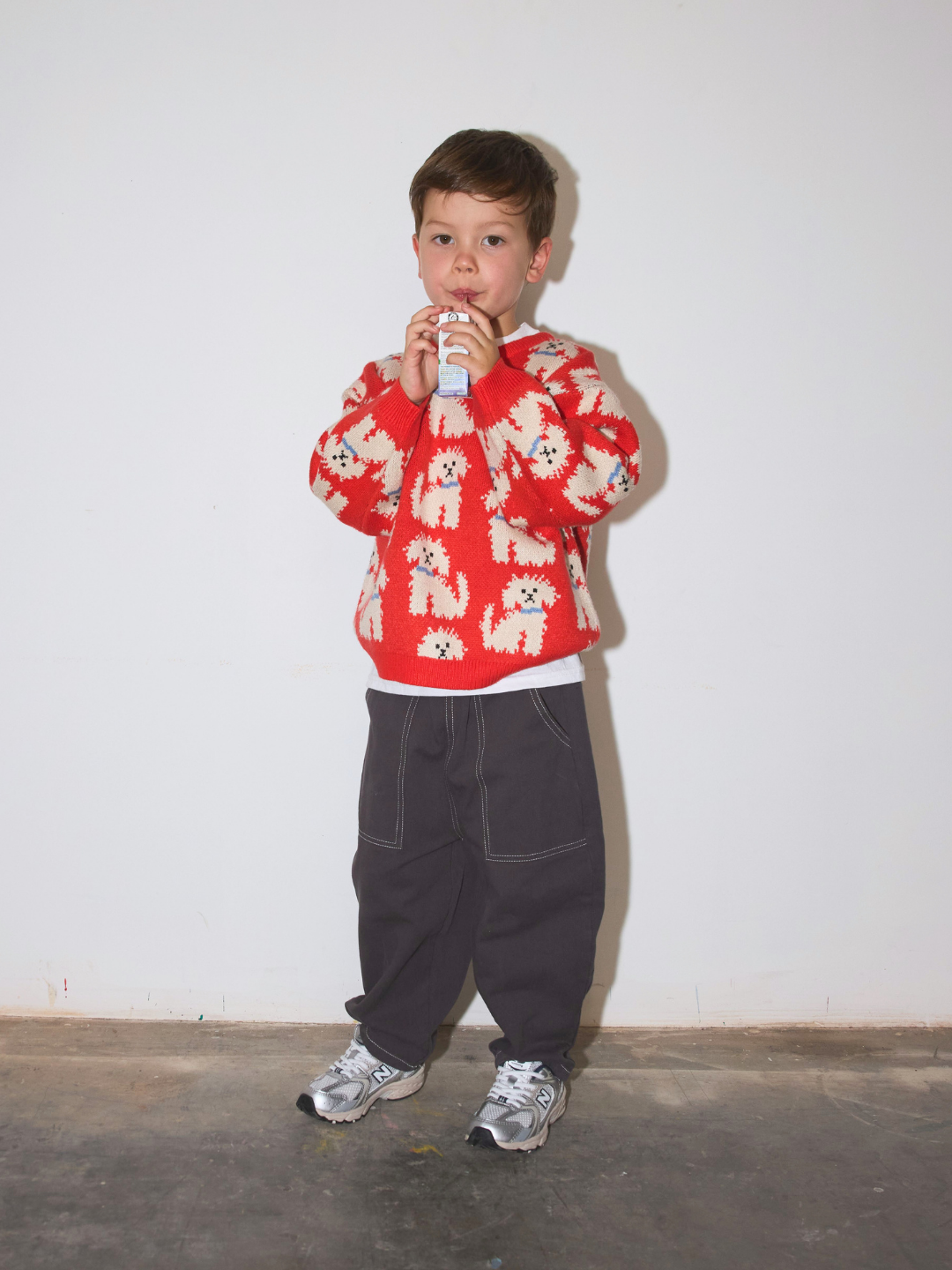 A young boy stands against a plain white wall wearing an orange knit sweater with a white dog pattern, loose dark pants, and sneakers, holding a juice box to his mouth.