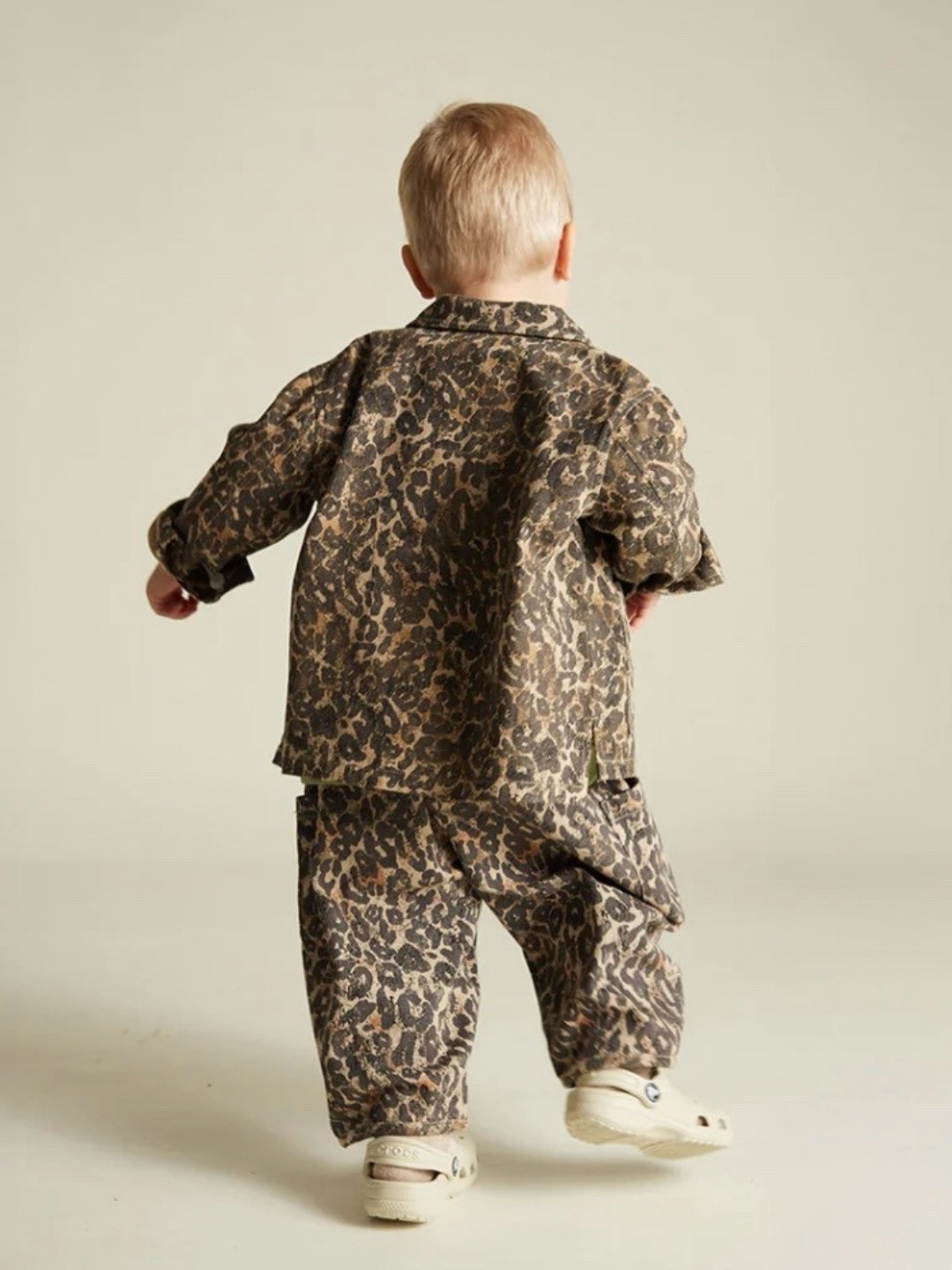 A toddler with short blonde hair wears a FEN LEOPARD JACKET made from organic cotton and white shoes, walking away on a light, plain background.