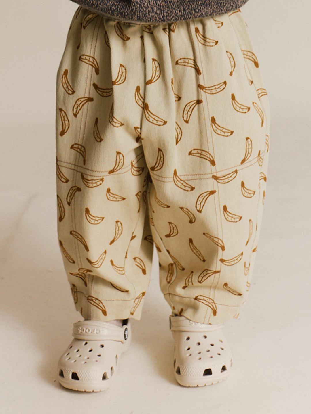 A child models the RIAN BANANA TROUSER, featuring an oversized balloon leg and banana print in organic cotton twill, paired with cream slip-on sandals on a light surface.