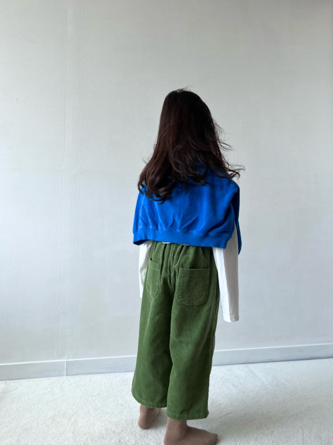 Moss | A girl wearing kids wide-leg corduroy pants in moss green, paired with a white t-shirt with three stripes in pink, red and green. A royal blue sweater is draped over her shoulders. She is standing on grey carpet with a white wall behind her, and facing backwards, showing the back view of the pants.
