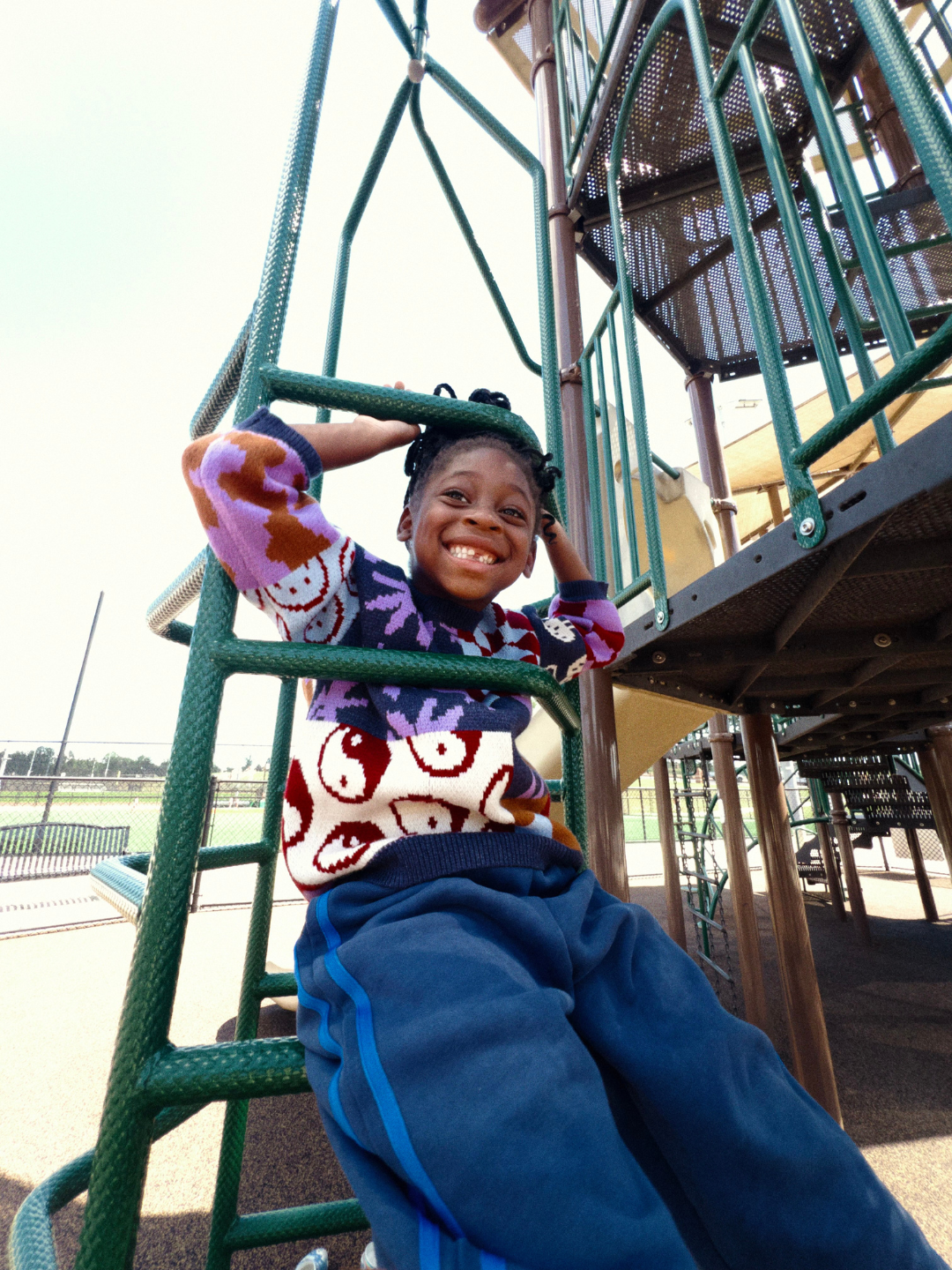 A child smiles while sitting on playground climbing equipment in the sun, wearing SUNDAY TRACKPANTS and a patterned sweater.