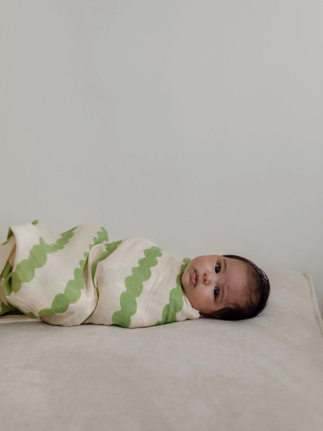 A baby wrapped in an ORGANIC BAMBOO SWADDLE WRAP with green and white hues lies on a light-colored surface, looking at the camera.