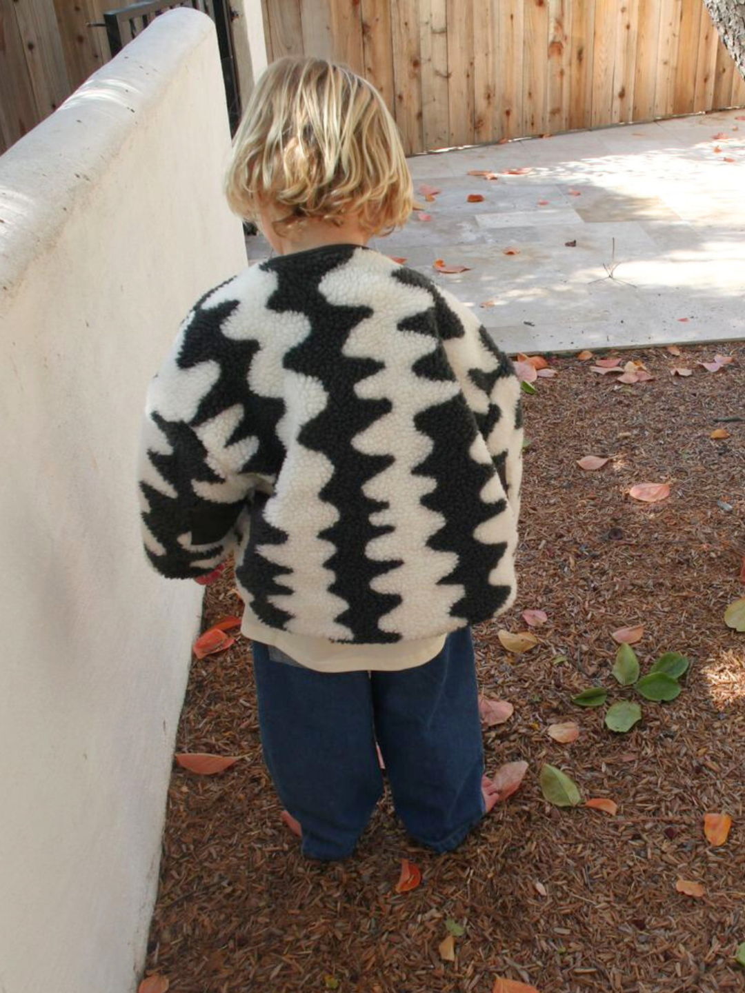 A child with blonde hair stands outdoors on a leaf-strewn ground, facing away from the camera, cozy in the WAVELENGTH SHERPA JACKET featuring a black and white pattern and quilted interior.