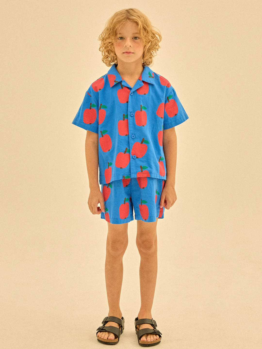 A child in the APPLE SUMMER SHIRT and shorts set, featuring red apple patterns, stands against a neutral background.