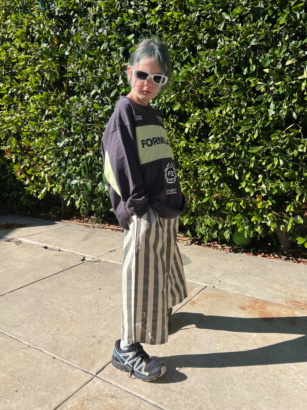 Person wearing a dark sweater with a visible brand logo, sunglasses, and a zebra-striped skirt standing against a green hedge.