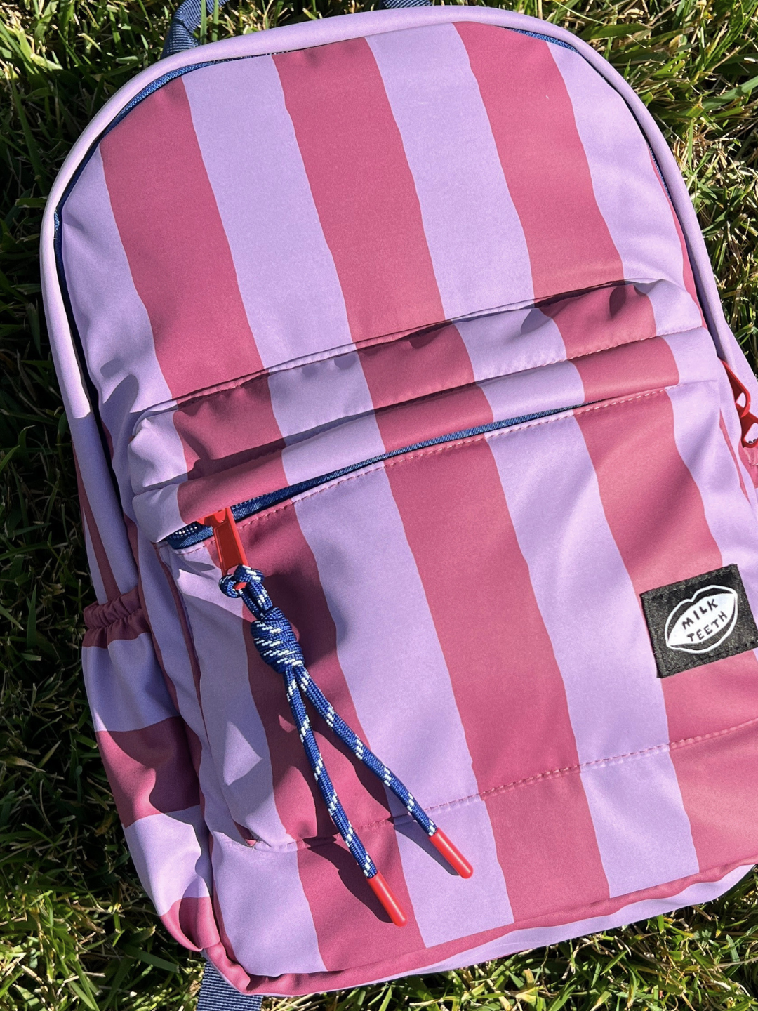 A kids backpack featuring vertical maroon and purple stripes, with a navy top handle, a front zippered pocket, and a black Milk Teeth logo patch on the pocket, shown lying on green grass.