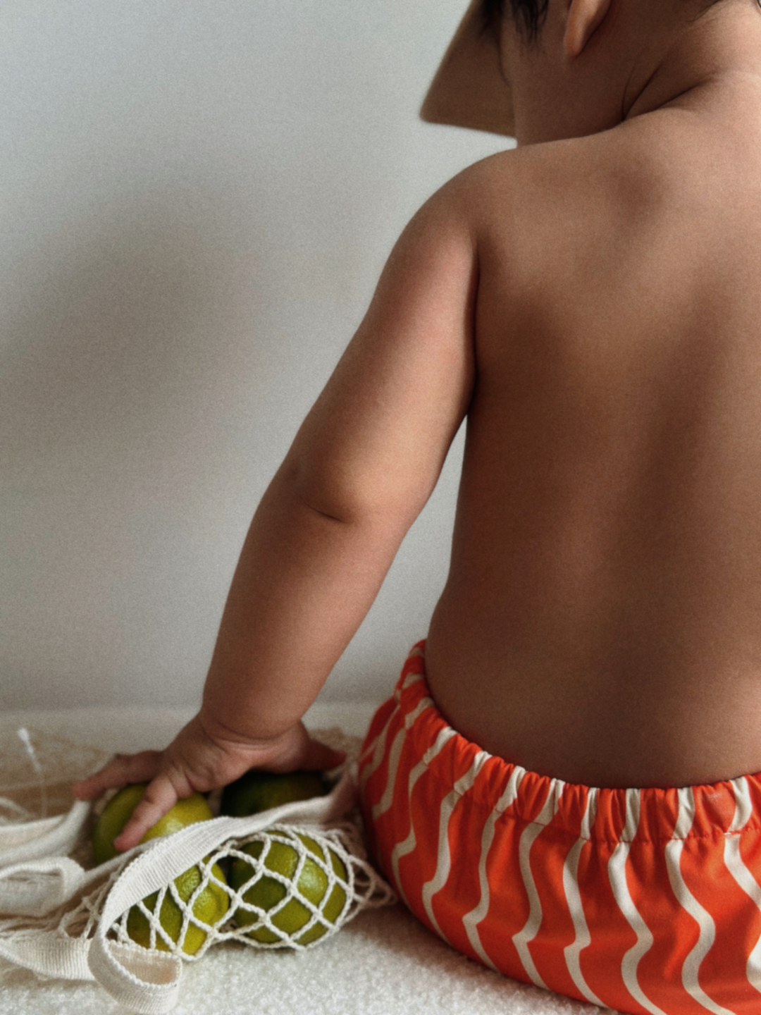 The back view of the swim diaper with an elastic waist with a tie and elastic leg holes on a child sitting down. The diaper is a bright orange with wavy and vertical cream colored lines.