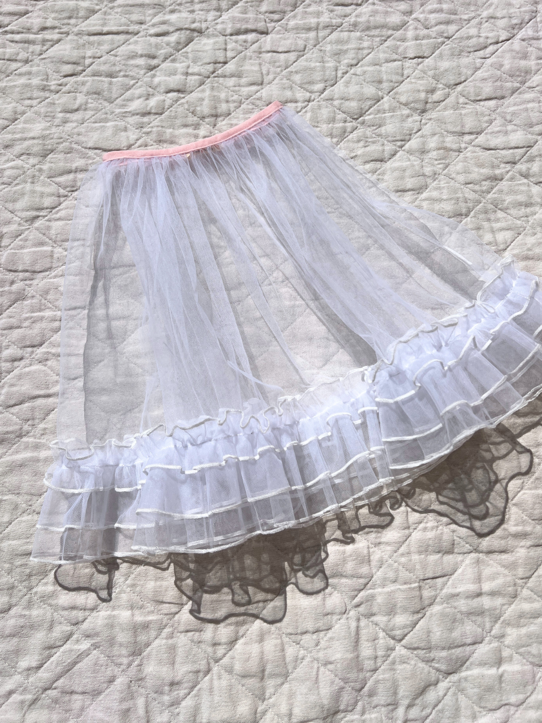 A front view of the kid's tulle layer skirt, white with a pink waistband, laying on an off-white cotton quilt.