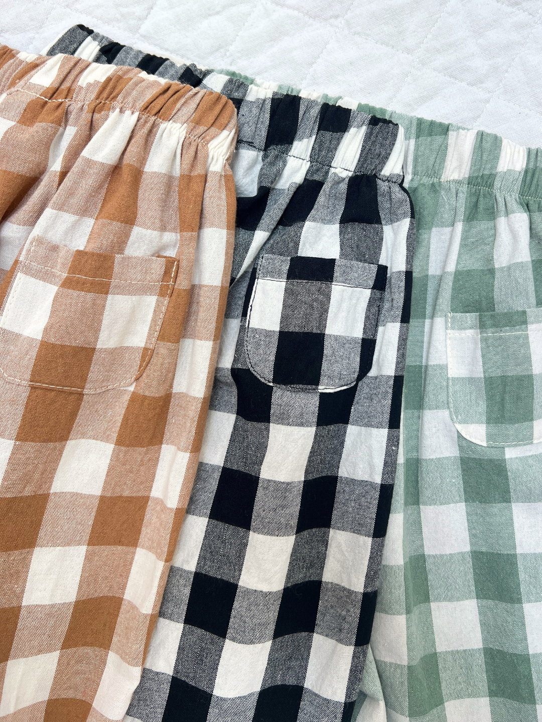 Brown + White | Three pairs of GINGHAM BABY PANTS in brown, black, and green with checkered pattern are displayed side by side, highlighting their elastic waistbands and back pockets.