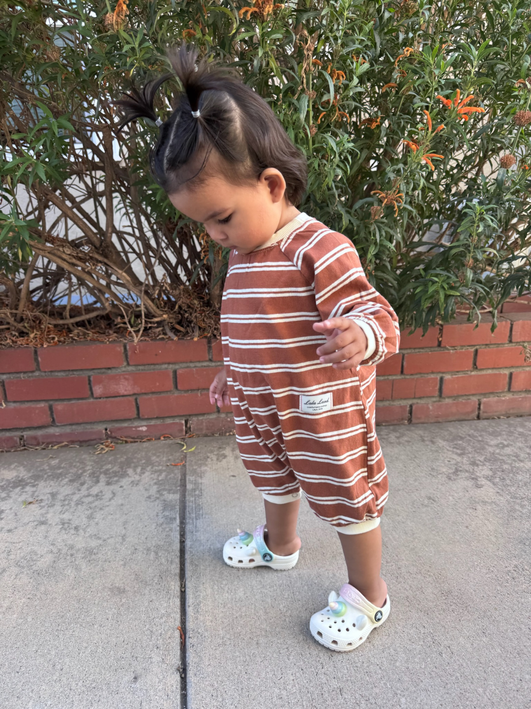 Terracotta | Child wearing a brown and white striped outfit standing on a sidewalk with greenery in the background