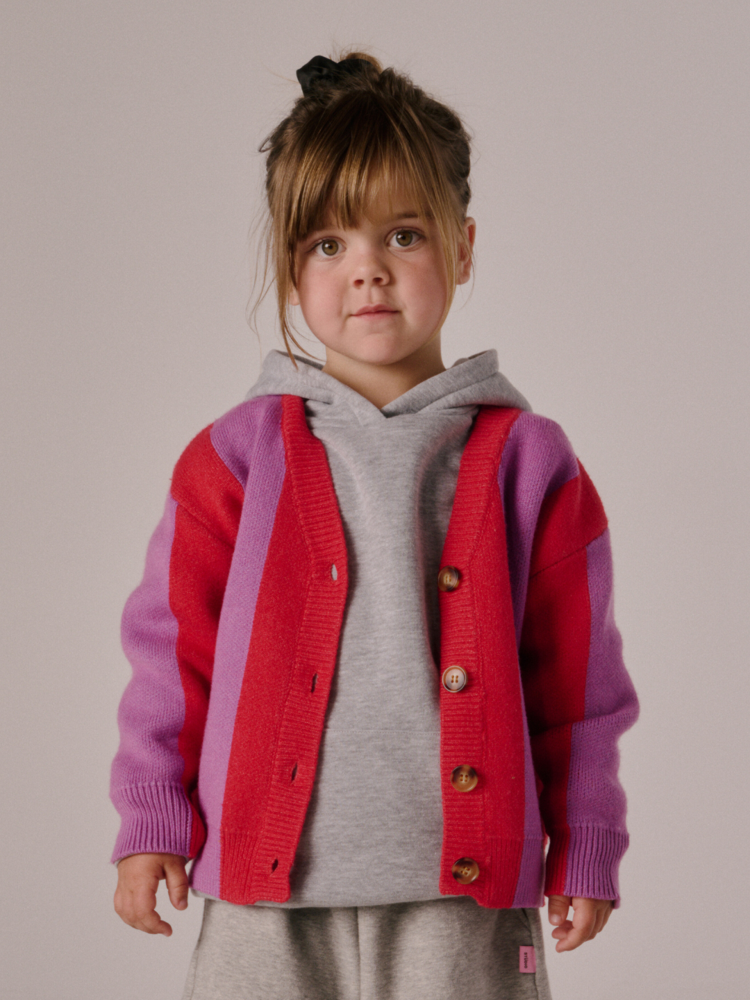 Red | A young child with brown hair in a ponytail wears a grey hoodie layered with the STRIPE CARDIGAN, featuring red and purple panels, standing against a plain background.