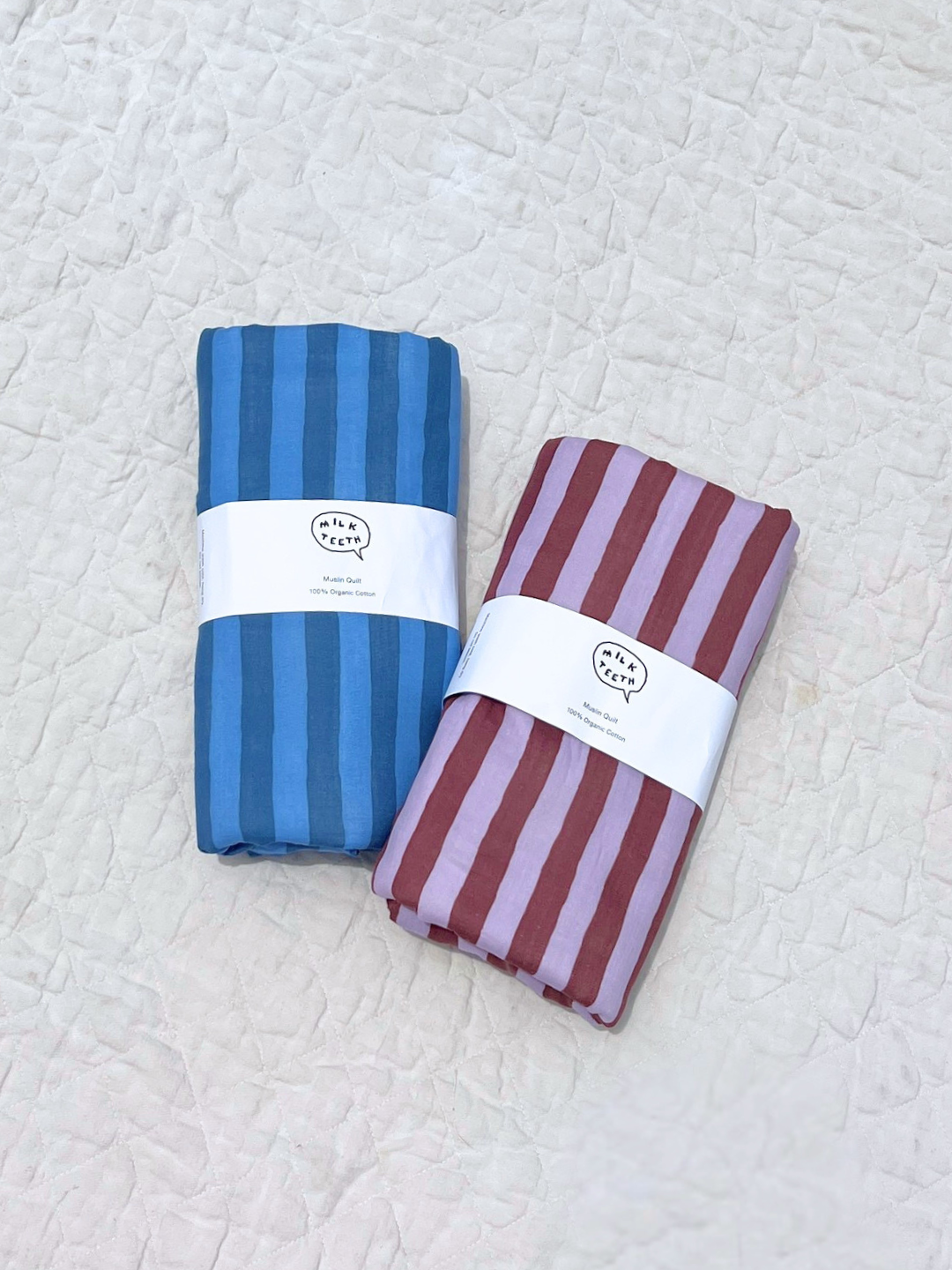  Two organic cotton quilts, one in blue stripes and one in purple and maroon stripes are shown rolled up with a white Milk Teeth label around the of each, and laid on a beige quilt.

