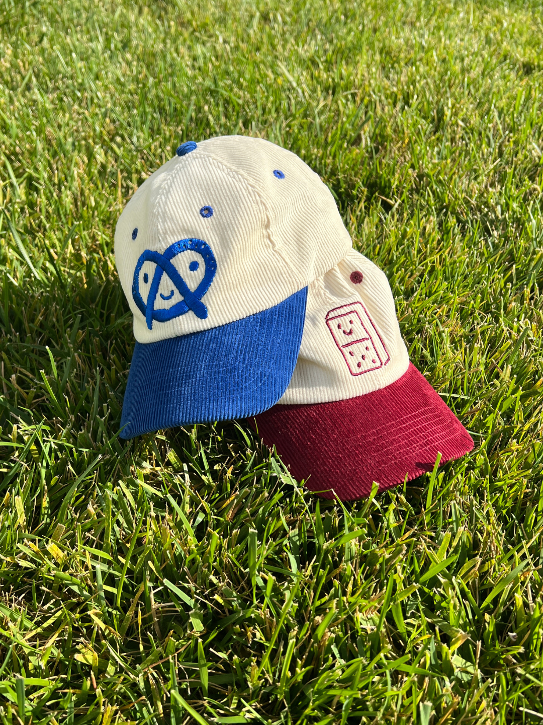 Two kids corduroy baseball caps resting on green grass. The caps features an off-white crown with a blue embroidered pretzel logo, a blue visor, and a maroon visor with a red embroidered domino icon.