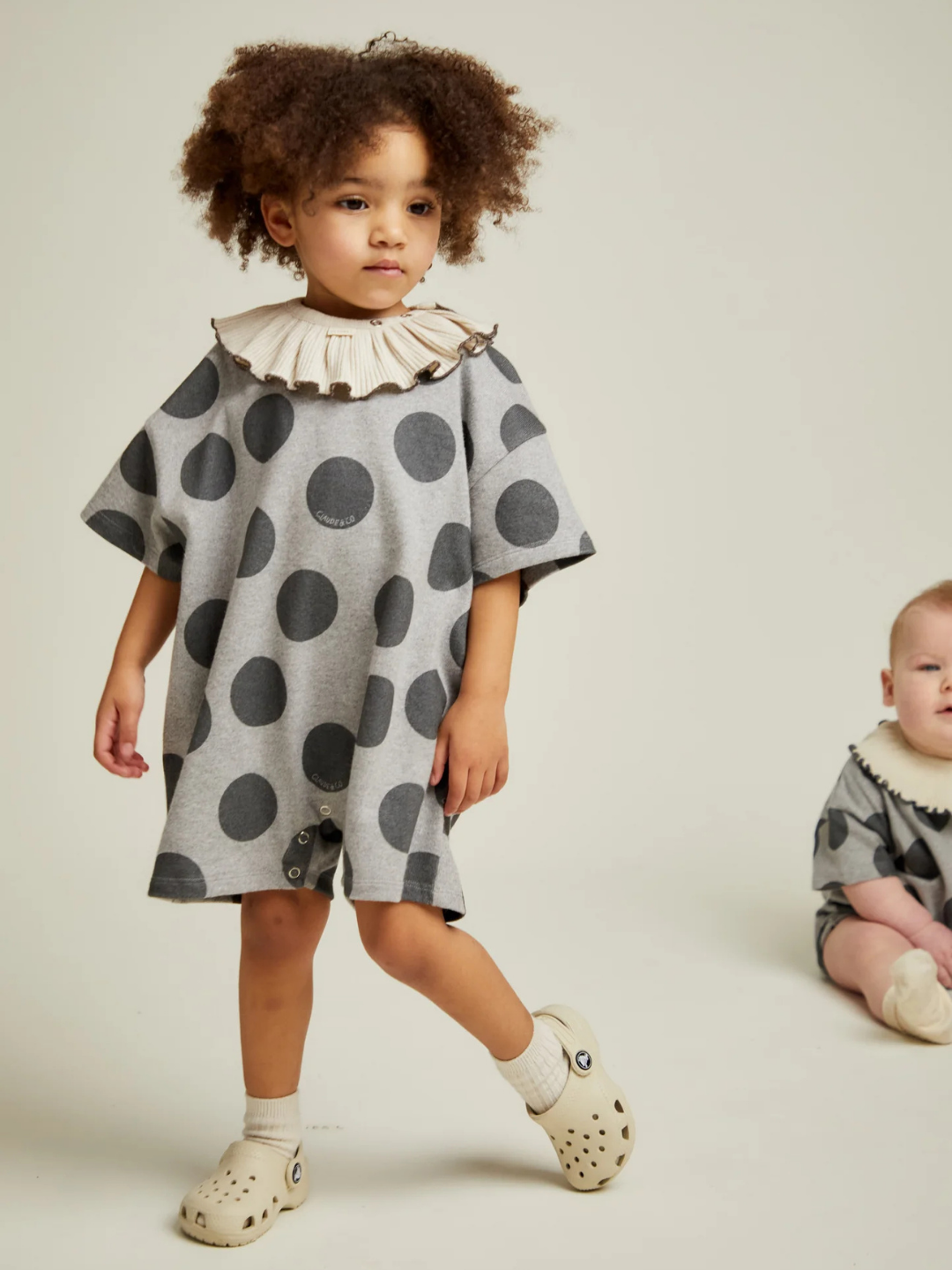 Two young children wear matching OPAL MARL SPOT ROMPERS with ruffled collars; one stands while the other sits on the floor against a plain background.