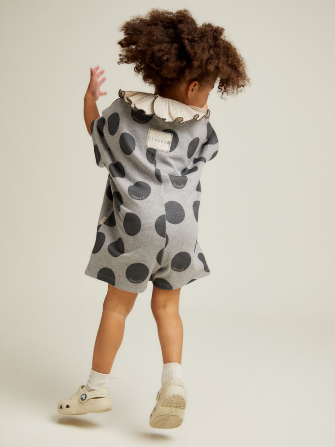 A young child with curly hair wears the OPAL MARL SPOT ROMPER—gray with bold black polka dots—paired with white socks and shoes, facing away from the camera against a plain background.