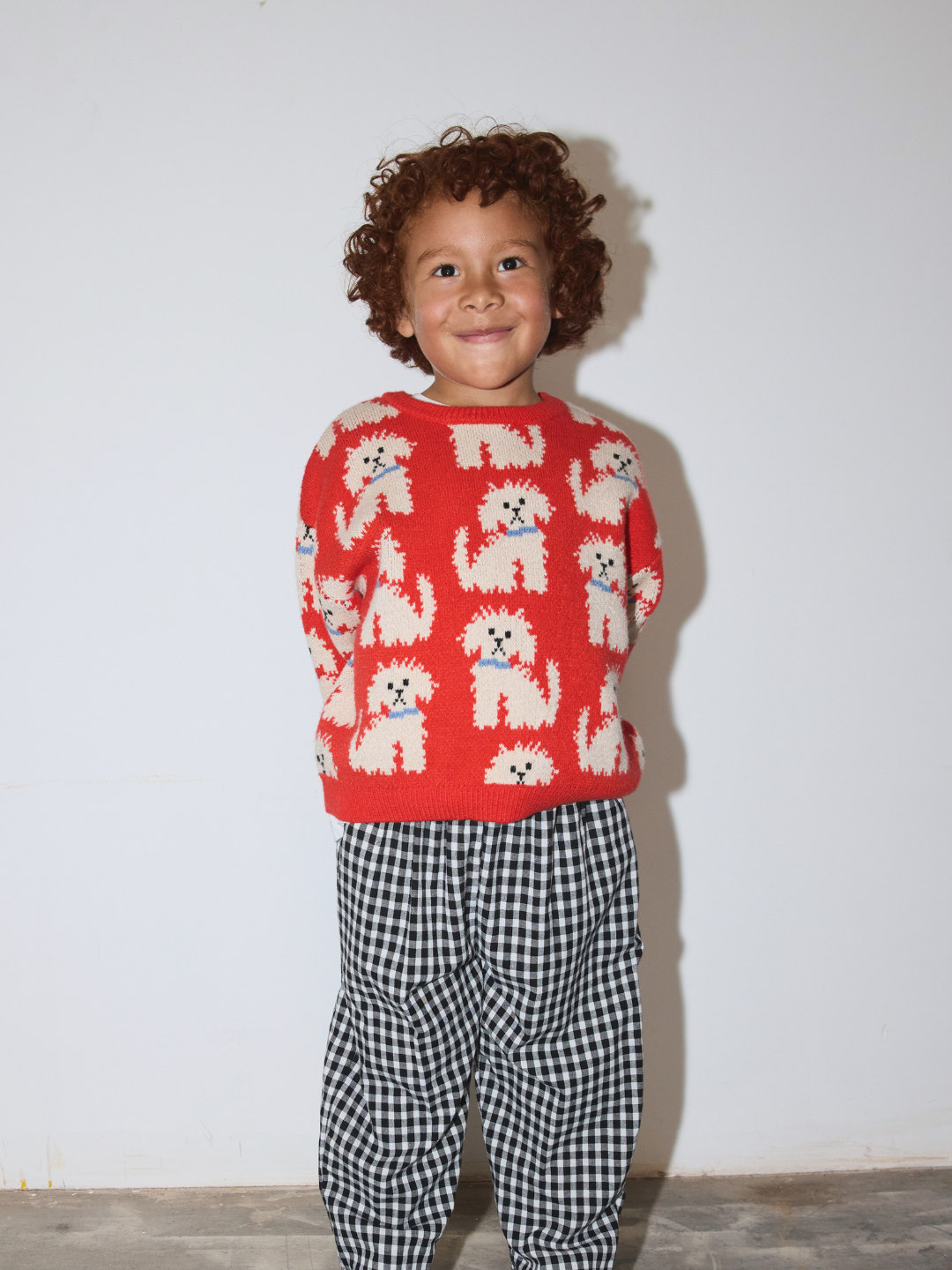 A young child with curly brown hair stands against a plain white wall wearing a bright orange knit sweater with a repeating pattern of small white dogs and black-and-white gingham pants.