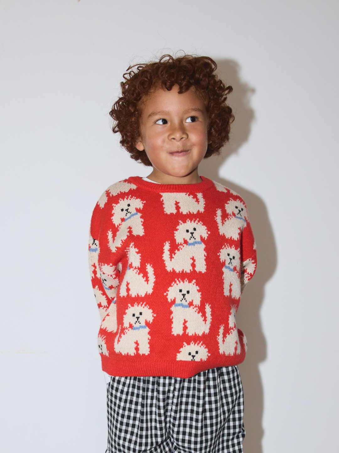 A young child with curly brown hair wearing an orange knit sweater with a repeating pattern of white poodles and black-and-white gingham pants, standing against a plain light gray background.