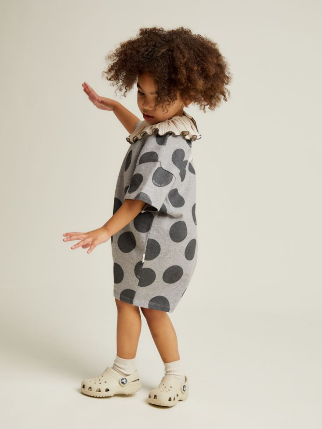 A young child with curly hair wears the OPAL MARL SPOT ROMPER, featuring a gray base with large black dots and a beige ruffled collar, paired with cream slip-on shoes, standing against a plain light background.