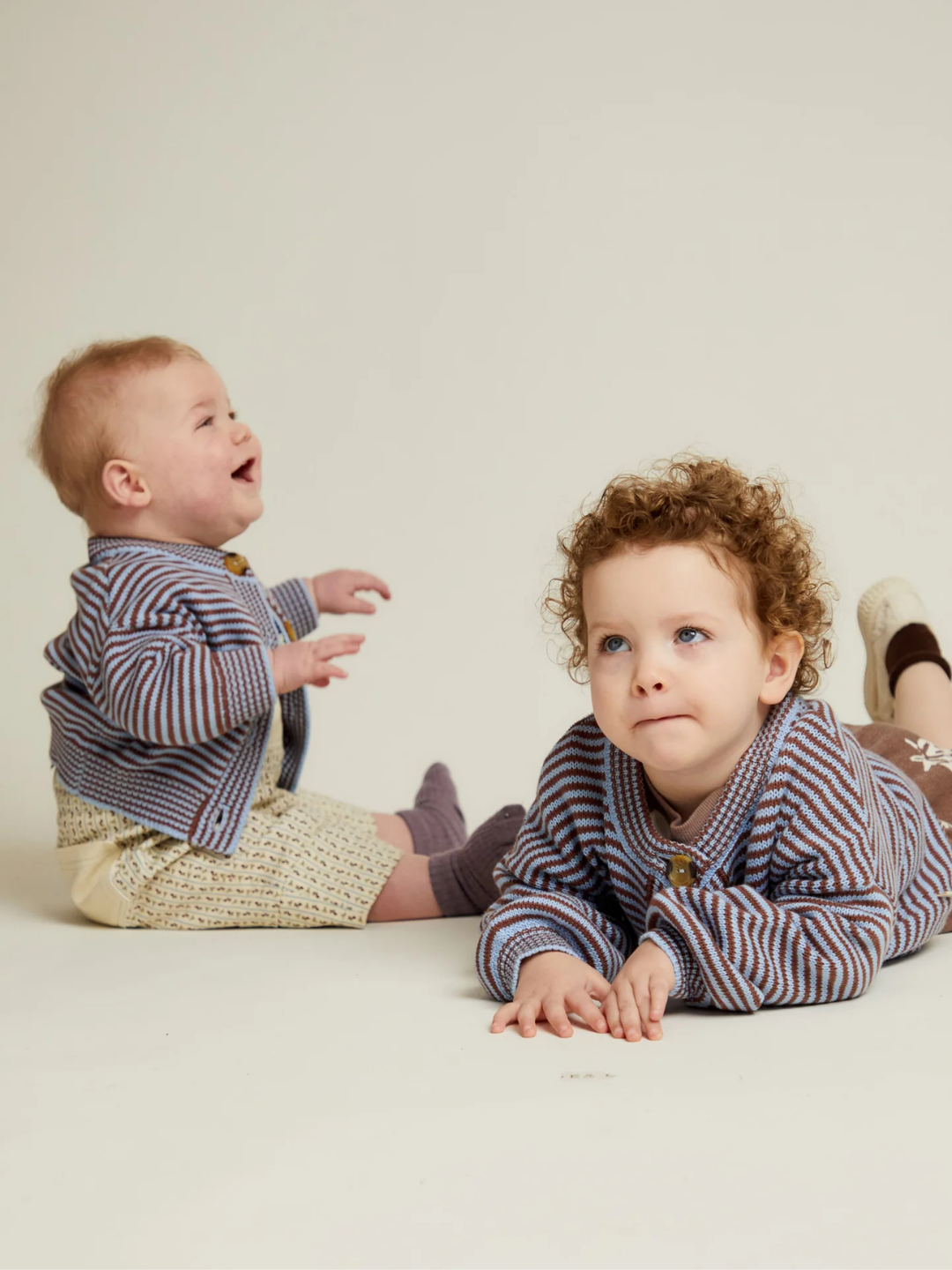 Two young children wear EMIL COCO SKY STRIPE CARDIGAN sweaters; one sits and smiles while the other lies on their stomach, both against a plain light background.