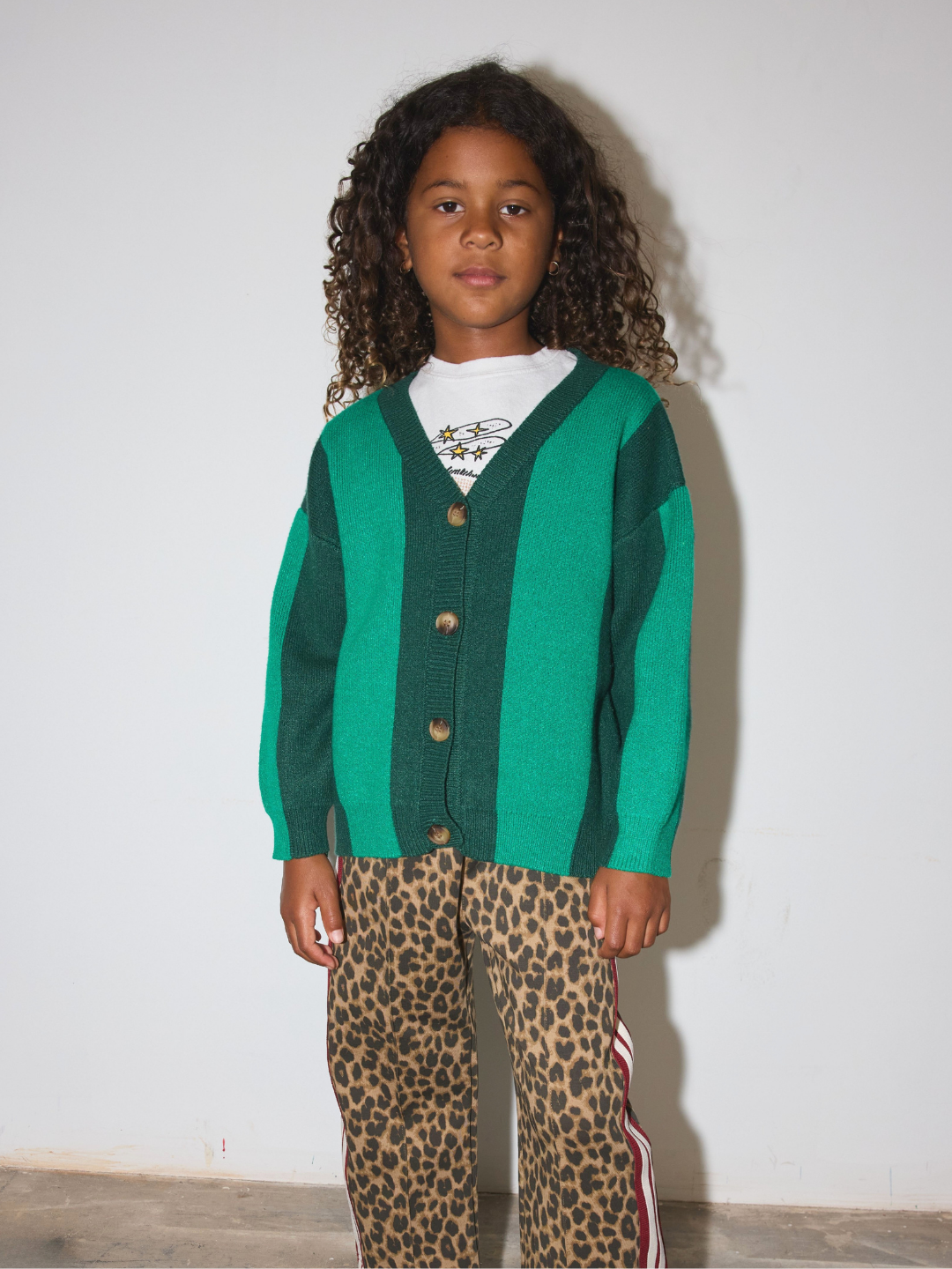 Green | A young girl with curly dark hair stands against a plain off-white wall wearing a green and dark green color-block knit cardigan over a white t-shirt, paired with leopard-print pants featuring a red and white stripe down the side.