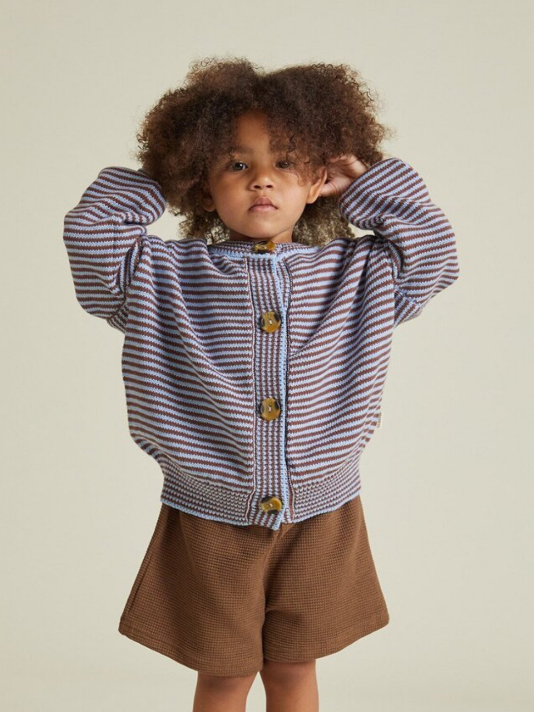 A young child with curly hair stands against a plain background, wearing the EMIL COCO SKY STRIPE CARDIGAN and brown shorts, holding their hands up to their head.