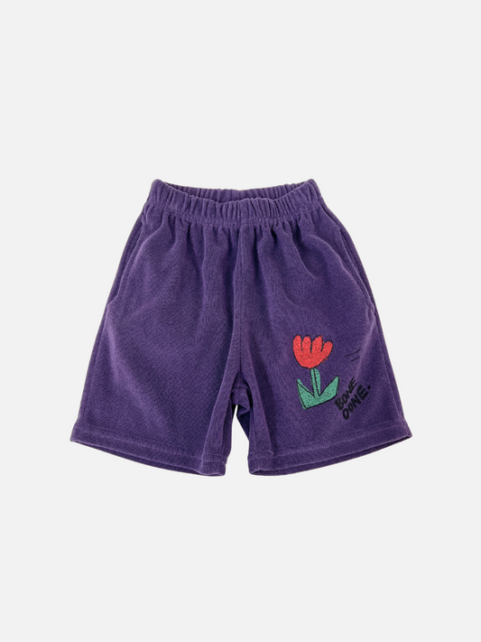 Image of The TULIP TERRY SHORTS are purple cotton shorts featuring an elastic waistband and a flower graphic near the hem. They offer a slouchy fit, ideal for casual days. This fun and comfortable style is from a popular Korean kids' brand.