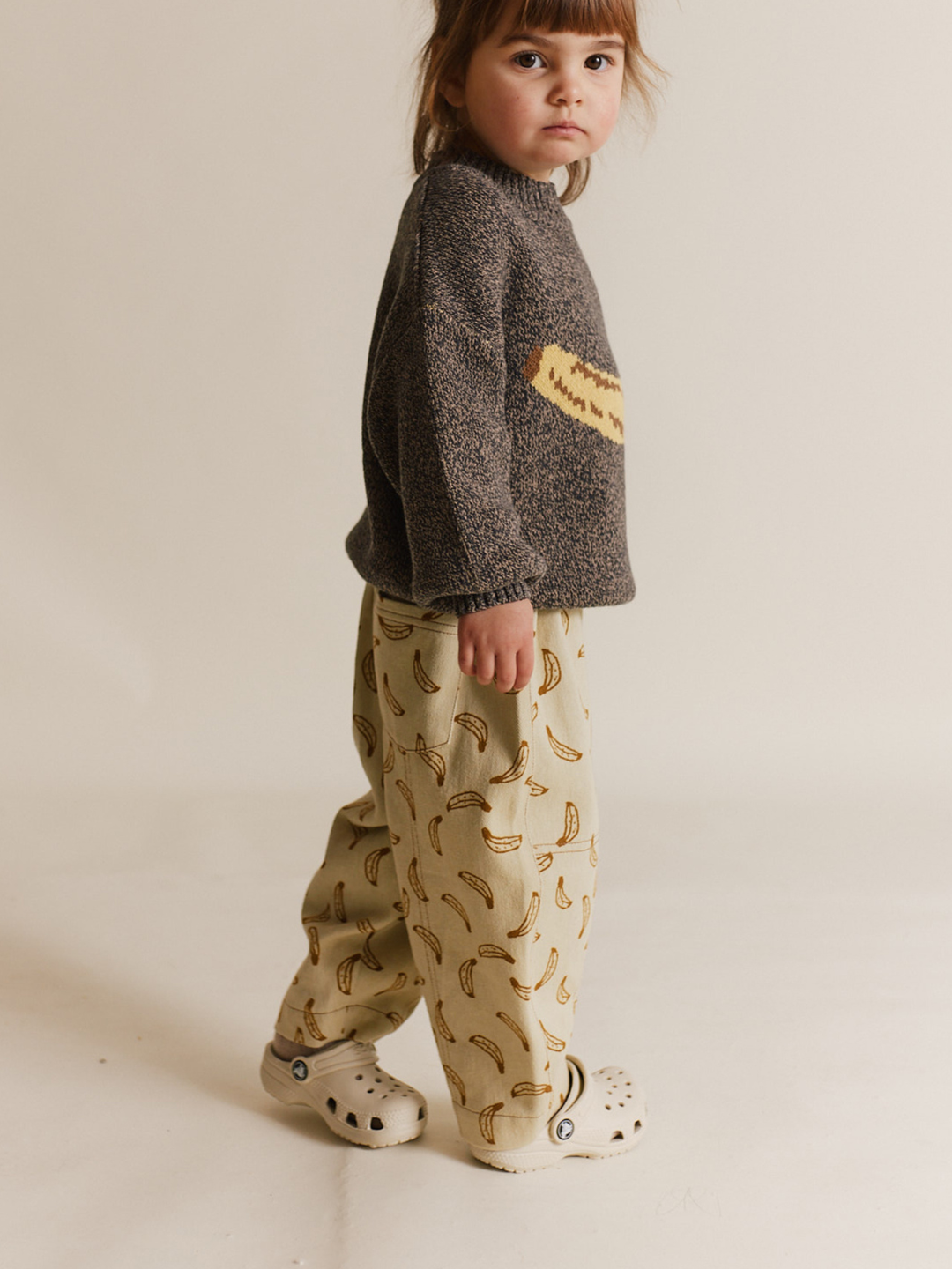 A young child stands sideways in RIAN BANANA TROUSER—oversized balloon-leg pants in organic cotton twill—paired with a grey sweater featuring a yellow design and light-colored slip-on shoes, set against a plain background.