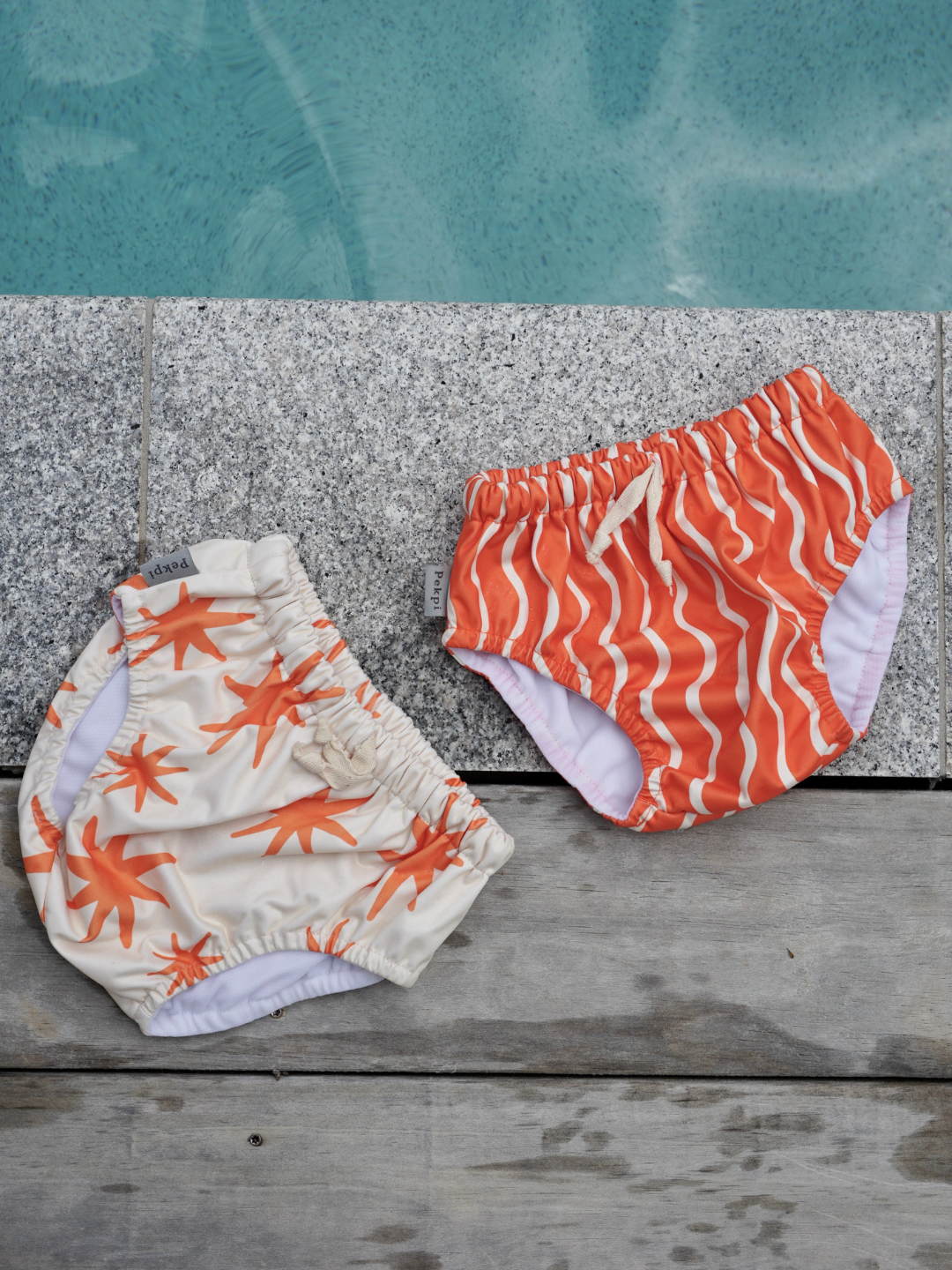 The front view of the swim diaper with an elastic waist with a tie and elastic leg holes. The diaper is a bright orange with wavy and vertical cream colored lines. It is laid on the ground next a pool with the Capri Swim Diaper.