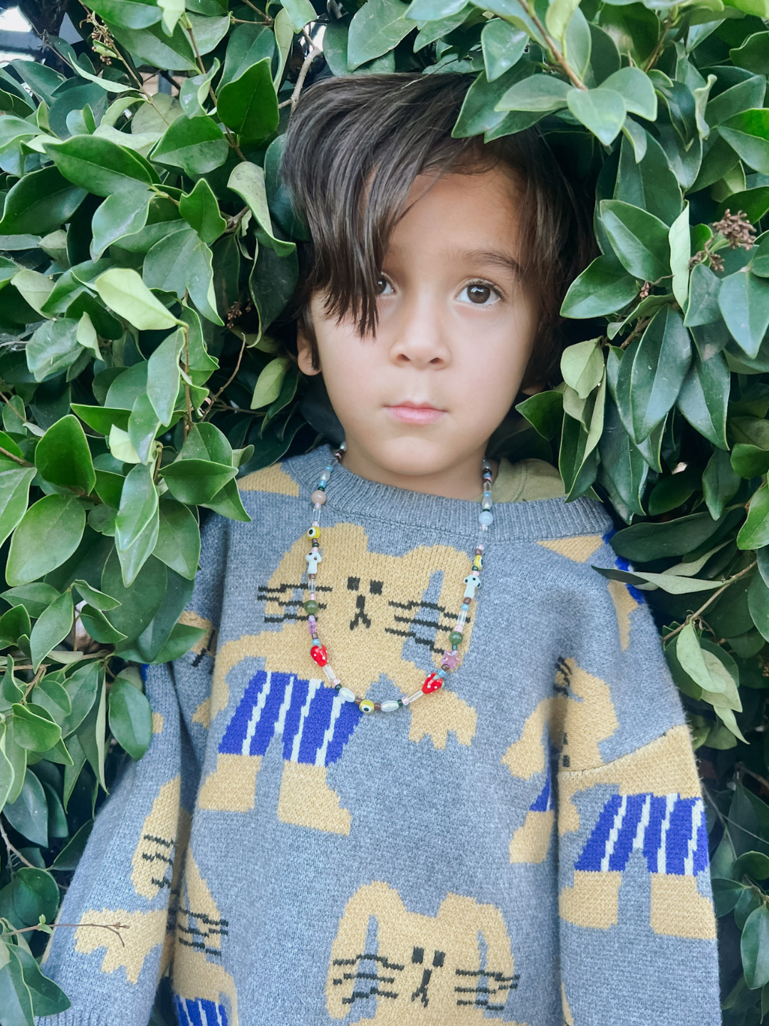 A young child with brown hair stands before green leaves, wearing a gray sweater with yellow cat designs and the GAIA NECKLACE—a genderless vintage glass bead necklace.