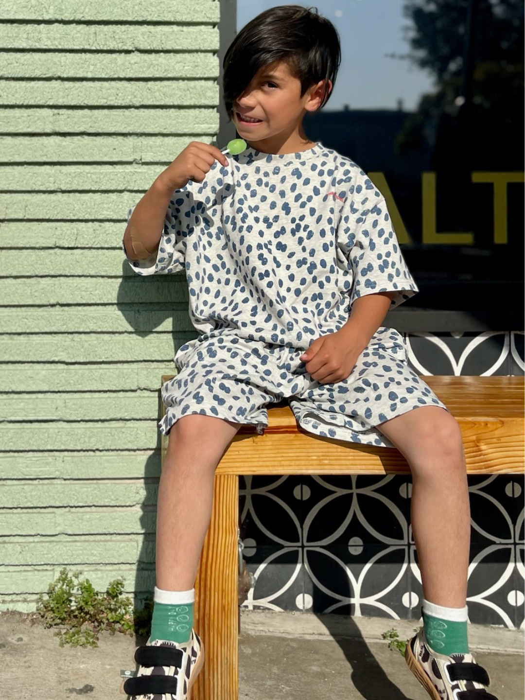 A young boy with dark hair wearing a relaxed SPOTTED TEE from a Korean kids brand sits on a wooden bench against a mint green wall. He holds a lollipop and has a playful smile. He wears green socks with patterns and black and white sandals. Sunlight creates a cheerful atmosphere.