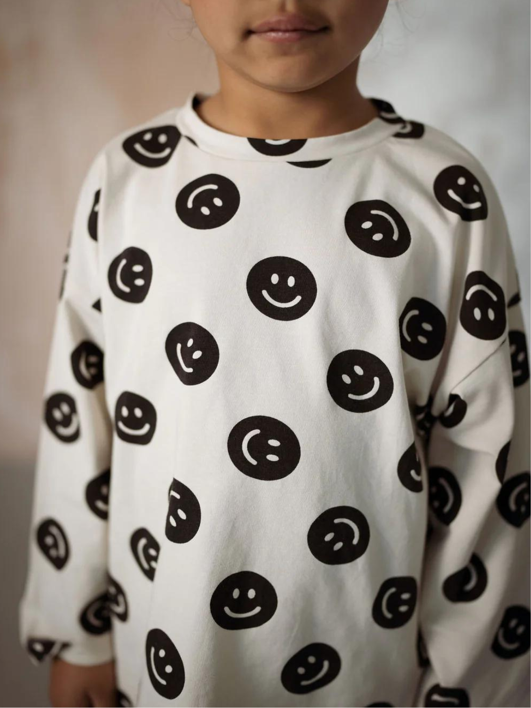 A zoomed in front view of the cream colored, long sleeve shirt with a black smiley face pattern on a child.