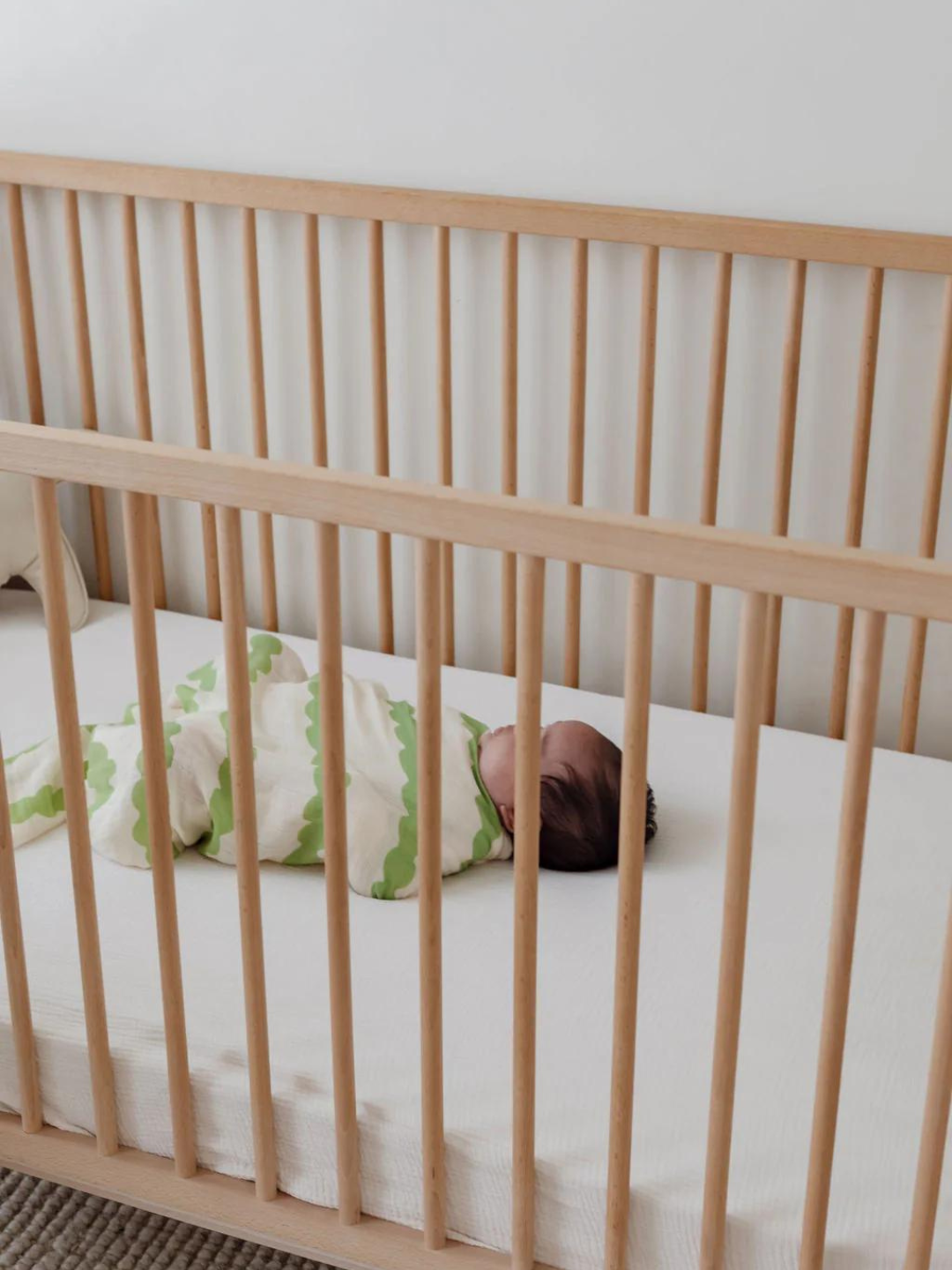 A baby wearing an Organic Bamboo Swaddle Wrap lies on its back in a wooden crib with vertical slats.