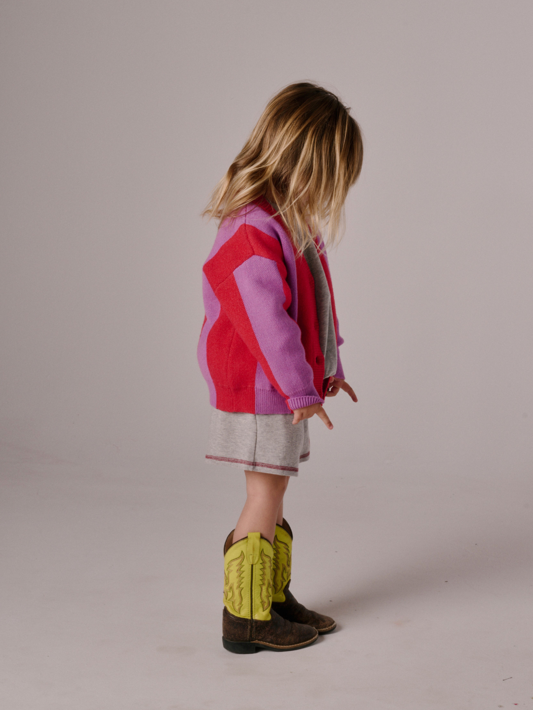 A young child with long blonde hair stands sideways in a STRIPE CARDIGAN, gray shorts, and yellow cowboy boots against a plain background.