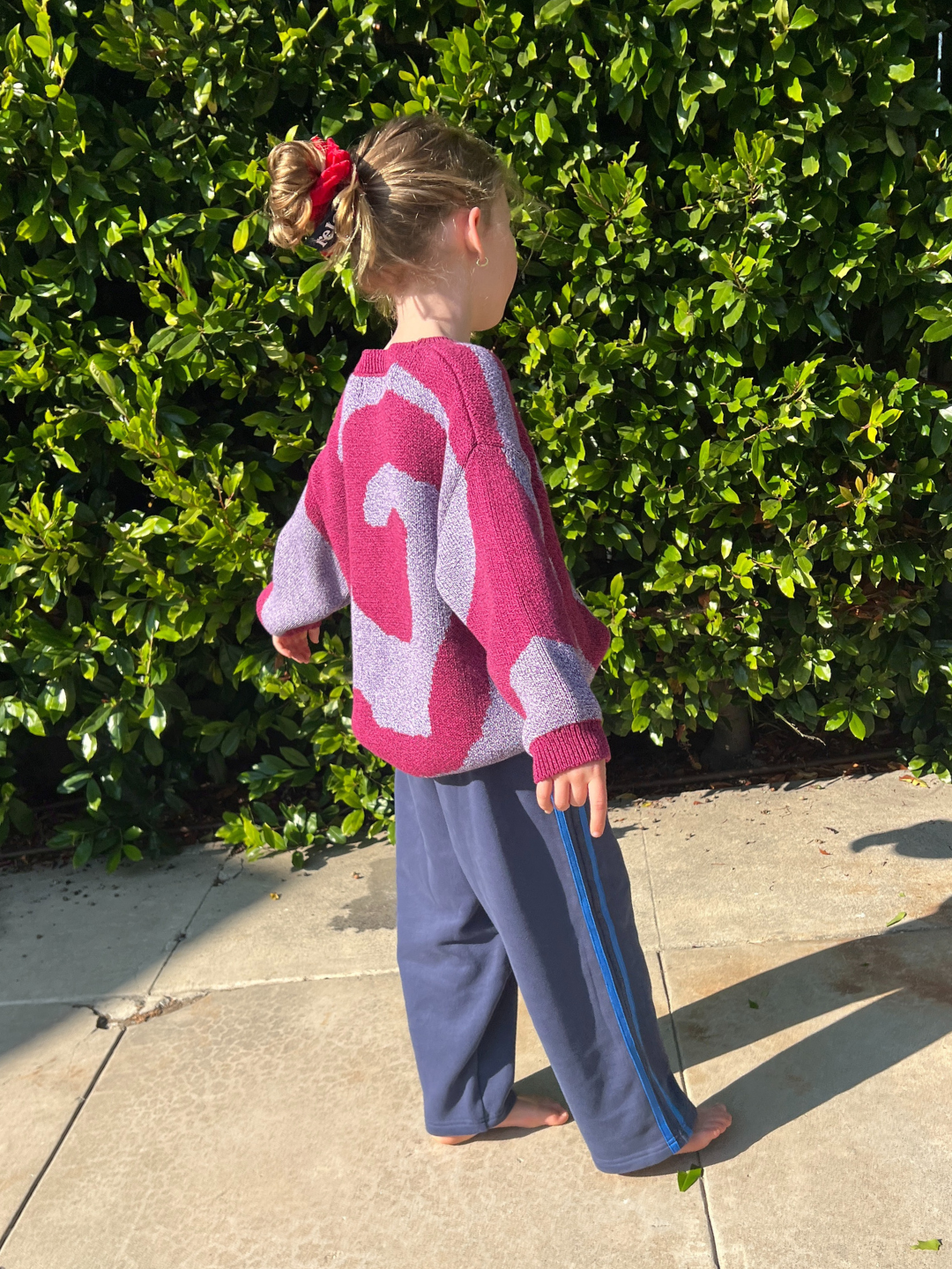 Pink/Purple | A child wearing the SWIRL SWEATER stands on a sunlit sidewalk in front of a green leafy hedge, paired with blue pants and a red hair tie.