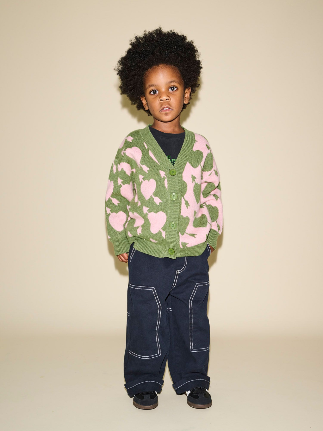 A young child with curly hair stands against a beige background, wearing the LOVESTRUCK CARDIGAN adorned with heart shapes in green and pink, paired with a black shirt, dark blue pants featuring white stitching, and black shoes. This vivid ensemble is perfect for capturing memorable photos.