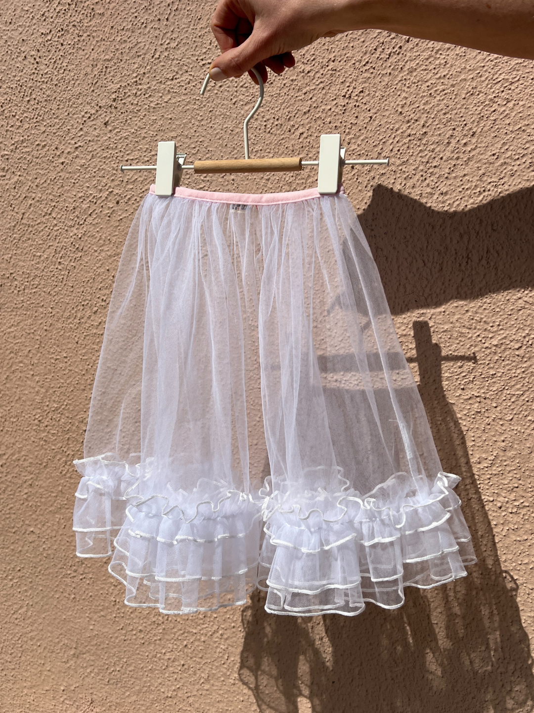 The kid's tulle layer skirt, white with a pink waistband, hanging on a white clip hanger, held by a hand against a peach colored textured wall, leaving a shadow.