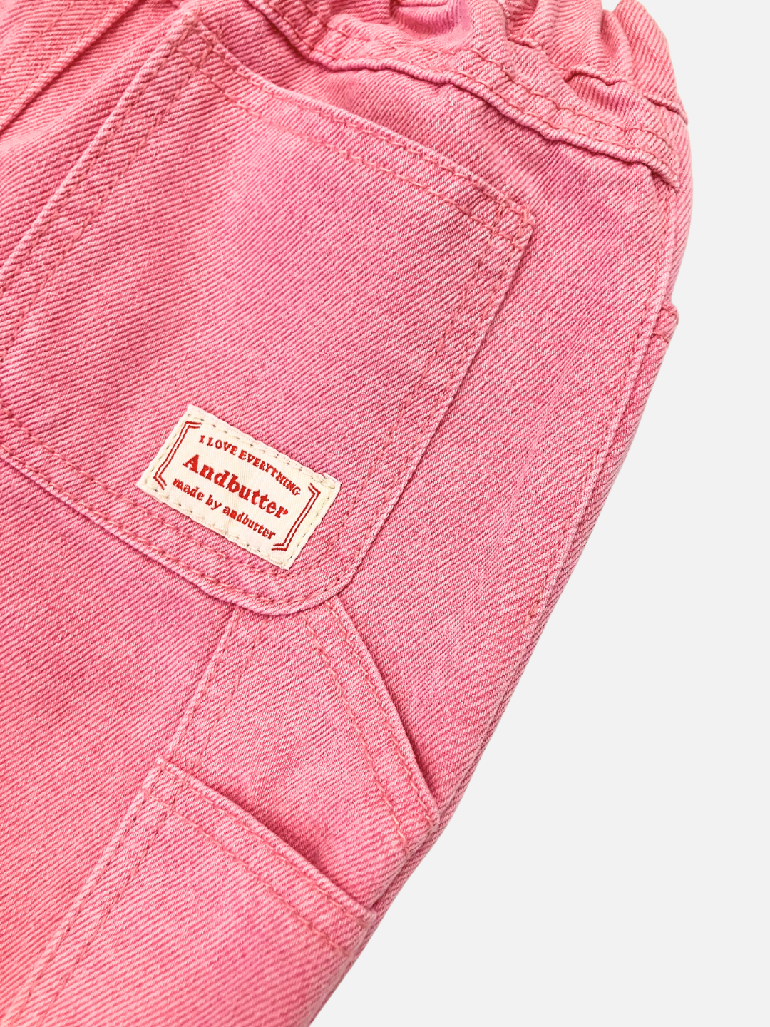 Peony | Close-up of the PIGMENT CARPENTER JEANS in pink, featuring pigment-dyed denim fabric with visible stitching and a front pocket. A small label reading "Andbutter" is sewn onto the fabric.