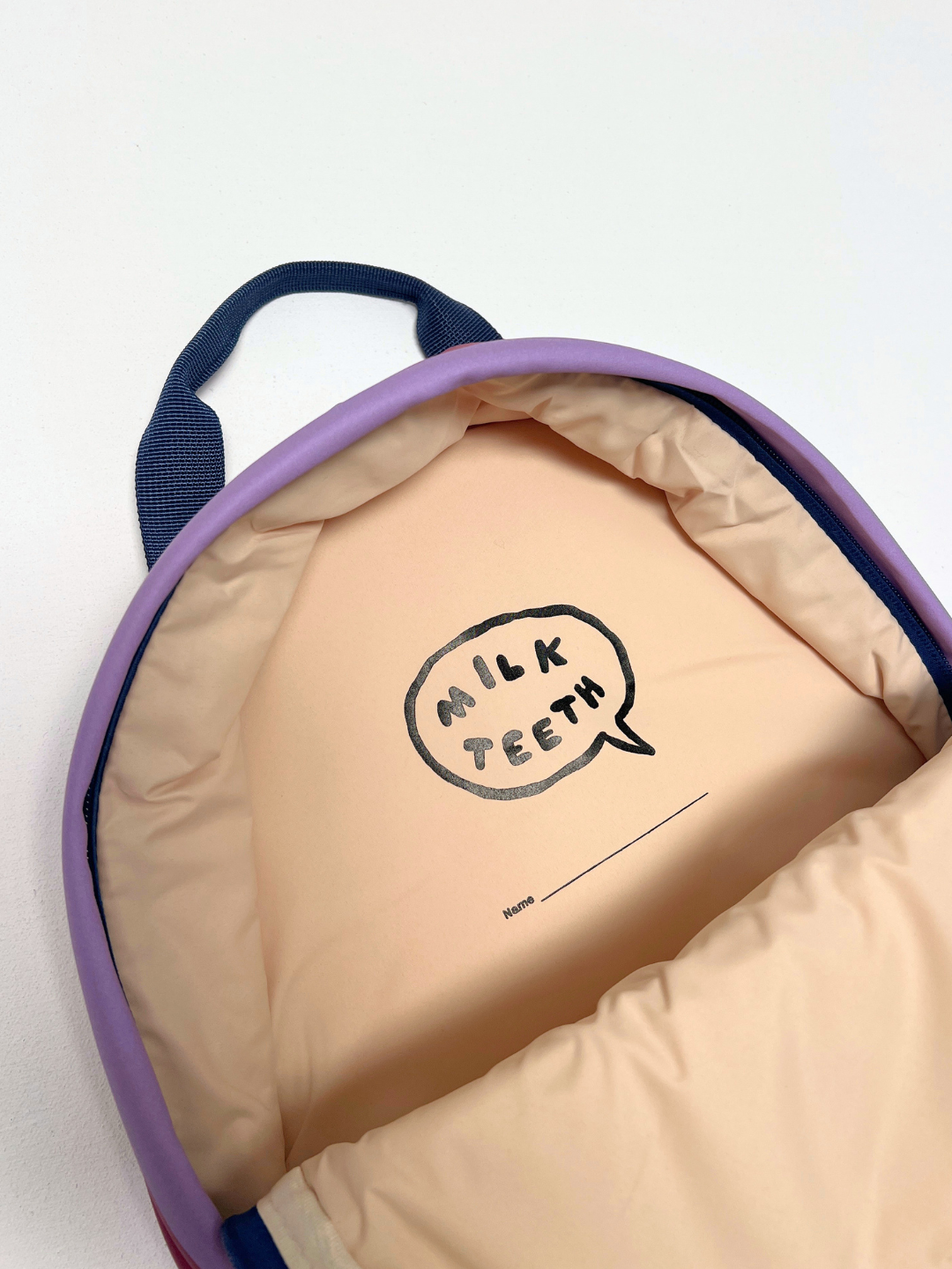 Purple Stripe | Close-up of the inside of a puffy quilted backpack with a beige interior, purple outer, navy top handle, and a printed speech-bubble logo reading 'MILK TEETH' on the lining with a name label space.