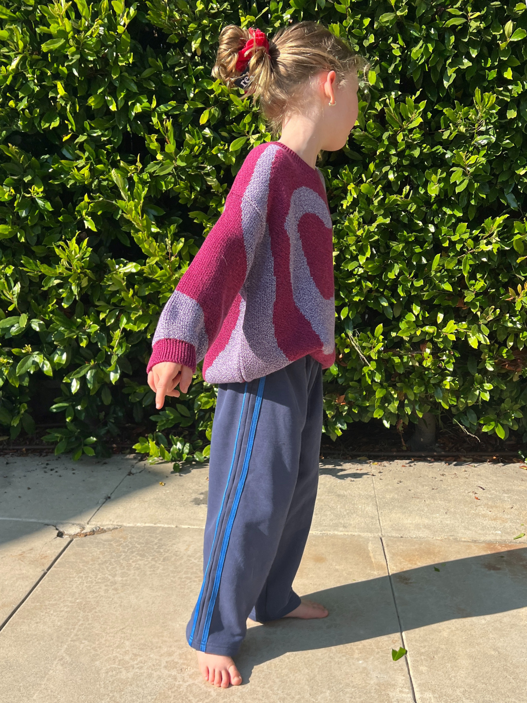 A young child, barefoot on sunlit pavement by green bushes, wears a pink and purple sweater with SUNDAY TRACKPANTS and a red hair tie.