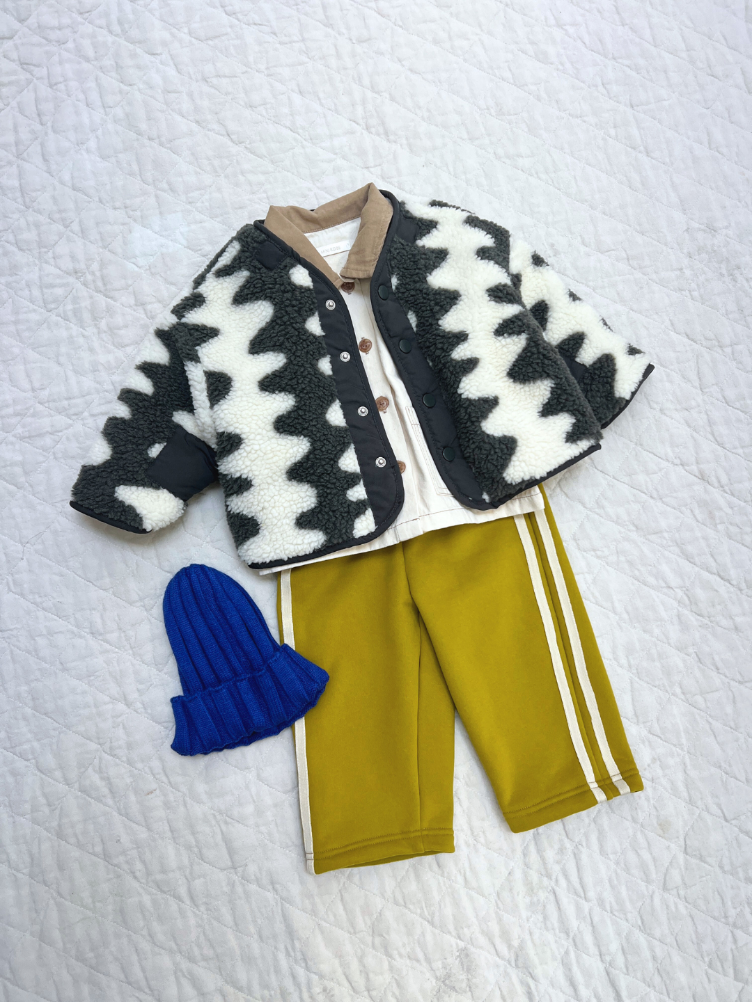 A children's outfit is displayed, featuring the WAVELENGTH SHERPA JACKET with a black and white zigzag pattern, a beige shirt, yellow striped pants, and a blue knit hat on a quilted surface.