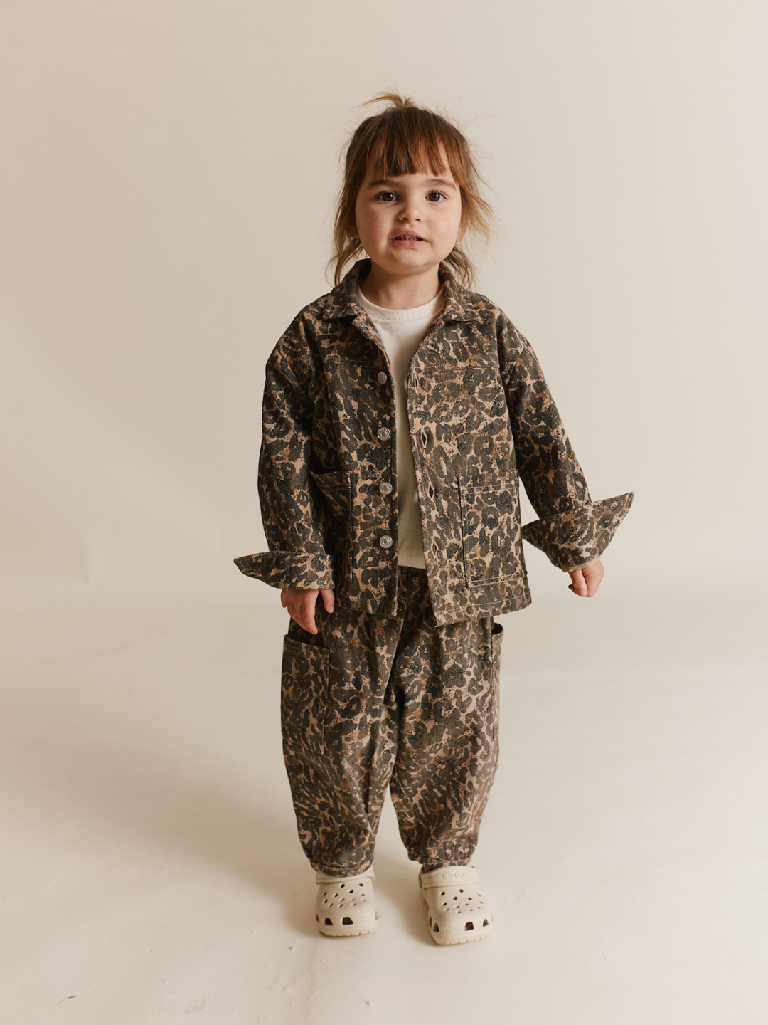 A young child in an oversized FEN LEOPARD JACKET and white clogs stands against a plain background, combining style with playful comfort.