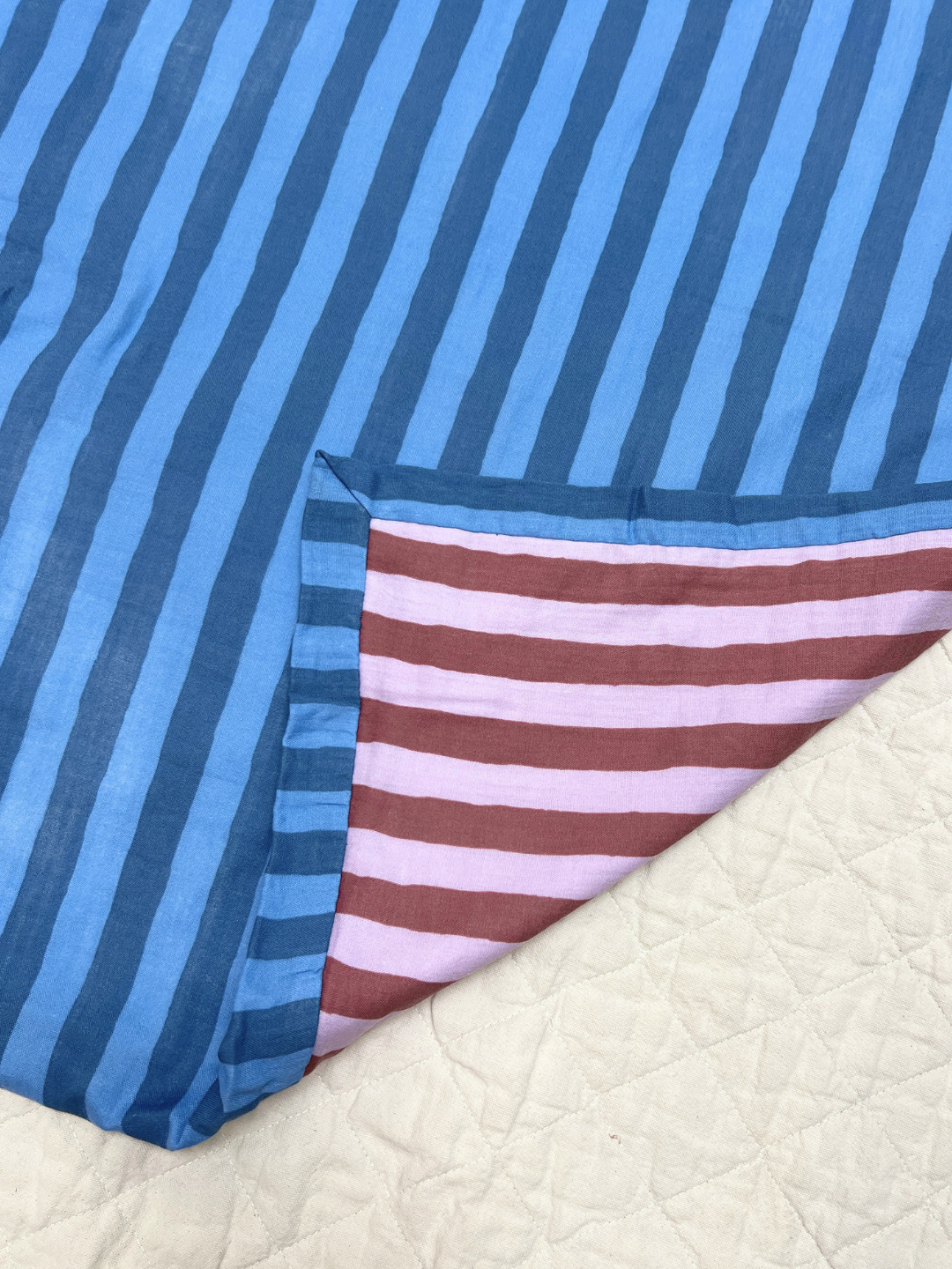 Detail shot of the organic cotton reversible in medium blue and darker blue stripes, with a corner folded back revealing its purple stripes underside, laid on a beige backdrop.