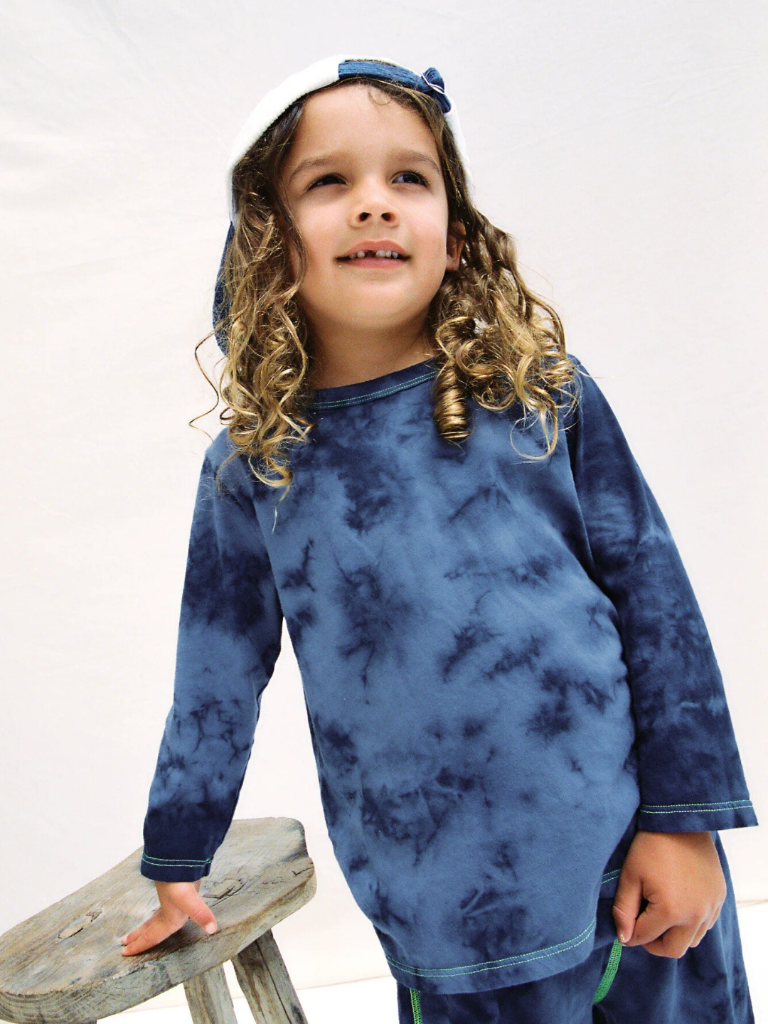Pacific Blue | Young child with long curly hair wears the COSMO TIE DYE LONG SLEEVE and matching hat, standing by a rustic wooden stool against a plain light background. The look channels laid-back Los Angeles style.