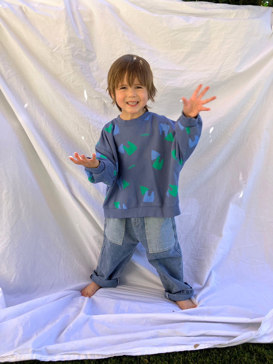 Child wearing the kids Duo Sweatshirt in faded blue printed with green and blue shapes and the word Navi. He is smiling and waving both hands, and wearing baggy blue jeans, and standing on a white sheet backdrop over grass.
