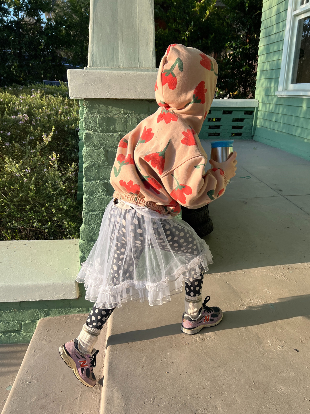 A child is wearing a tulle layer skirt that is see through over polka dot leggings, with a big printed hoodie , chunky socks and purple sneakers.