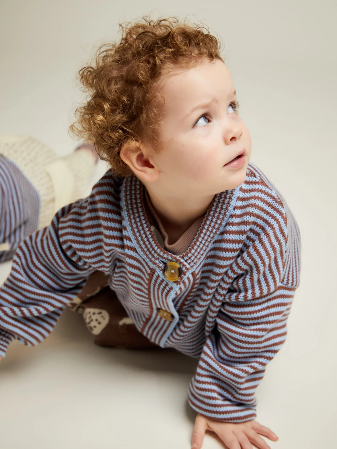 A young child with curly hair sits on the floor, looking upward, dressed in the EMIL COCO SKY STRIPE CARDIGAN.