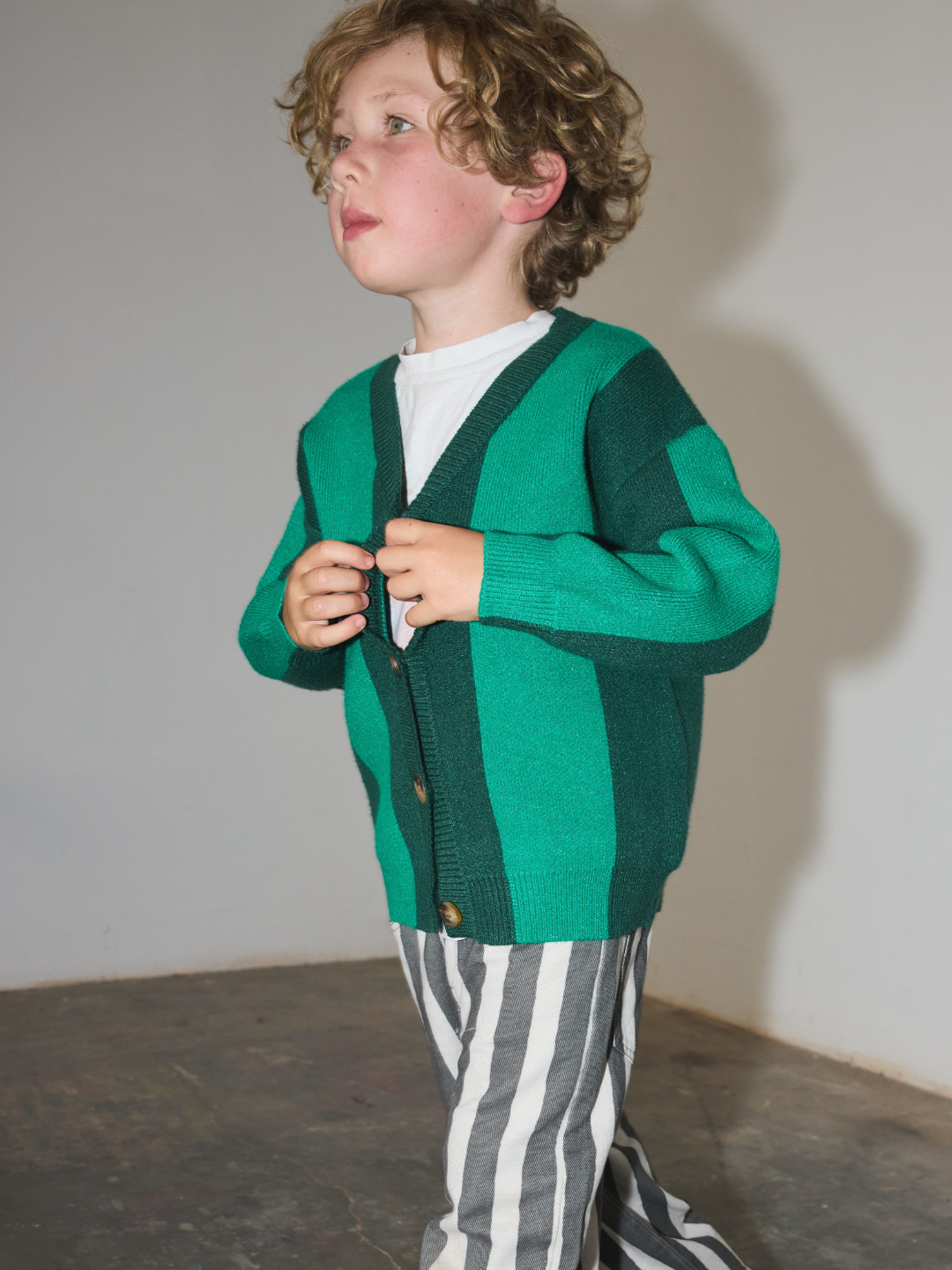 Green | A young child wearing a green and dark green striped knit cardigan over a white t-shirt and gray-and-white vertically striped pants, standing against a plain light gray wall.