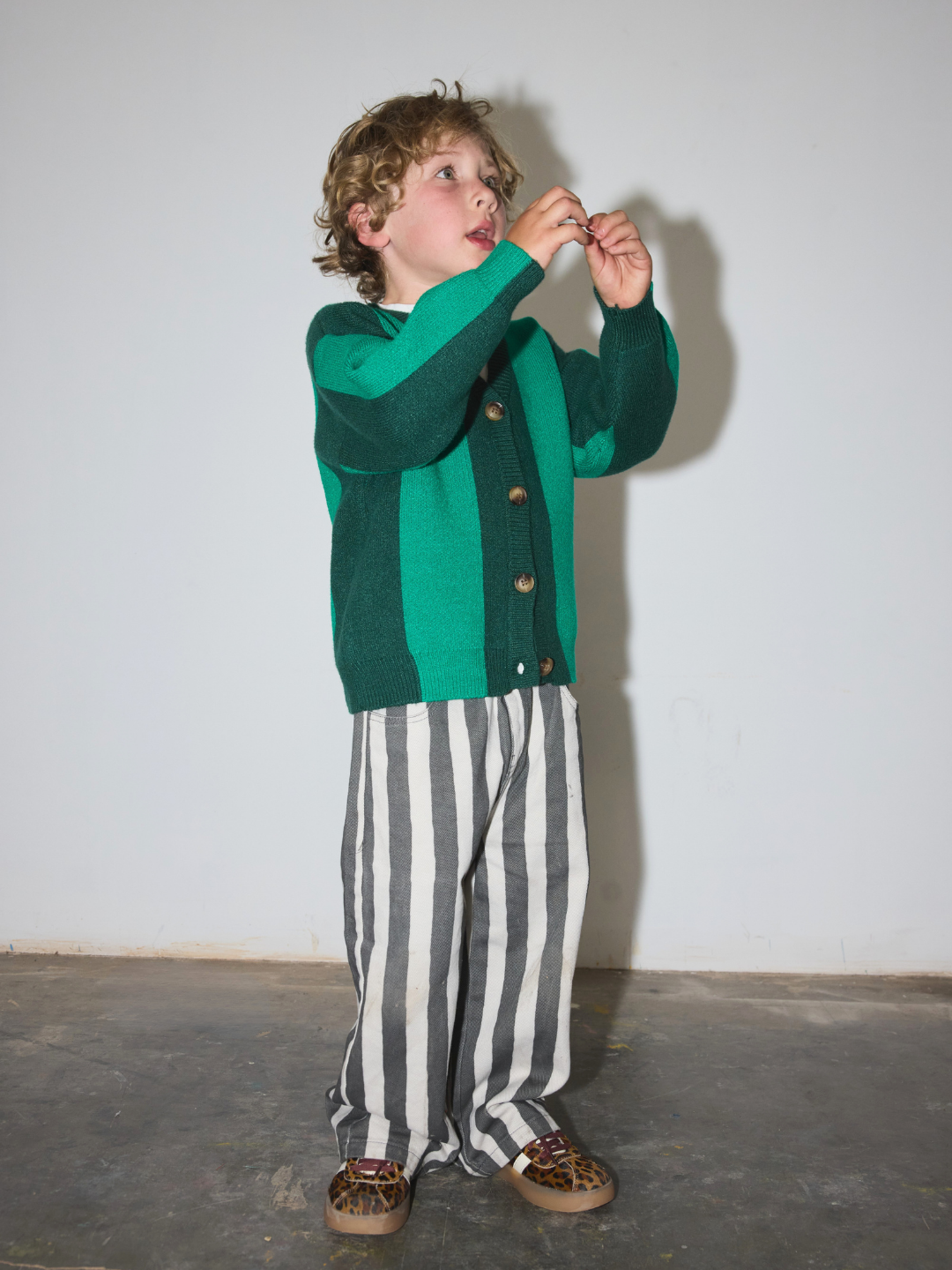 A child wearing a green and teal striped cardigan, black and white vertically striped trousers, and leopard print shoes, standing against a plain light gray wall.